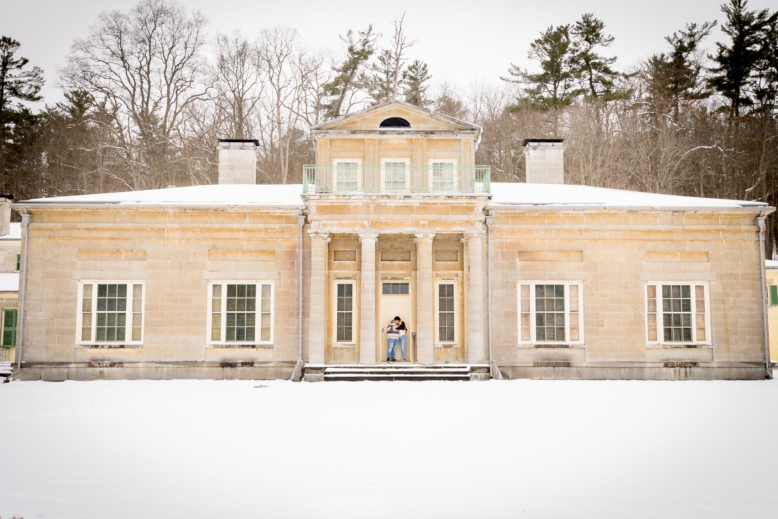 A couple stands at the entrance of a historic yellow stone mansion surrounded by winter snow.