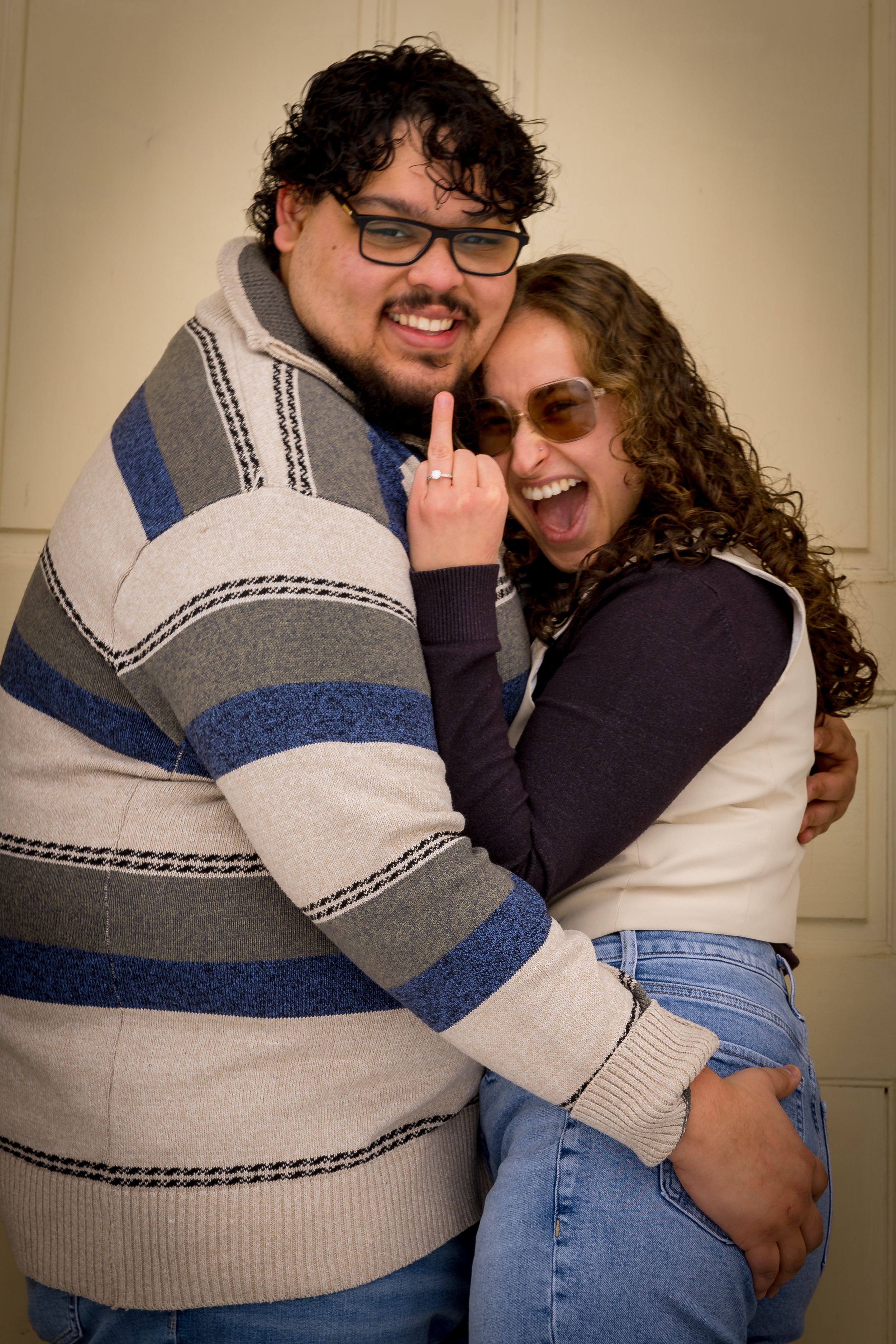 A laughing couple poses playfully together, with the woman showing off her engagement ring.