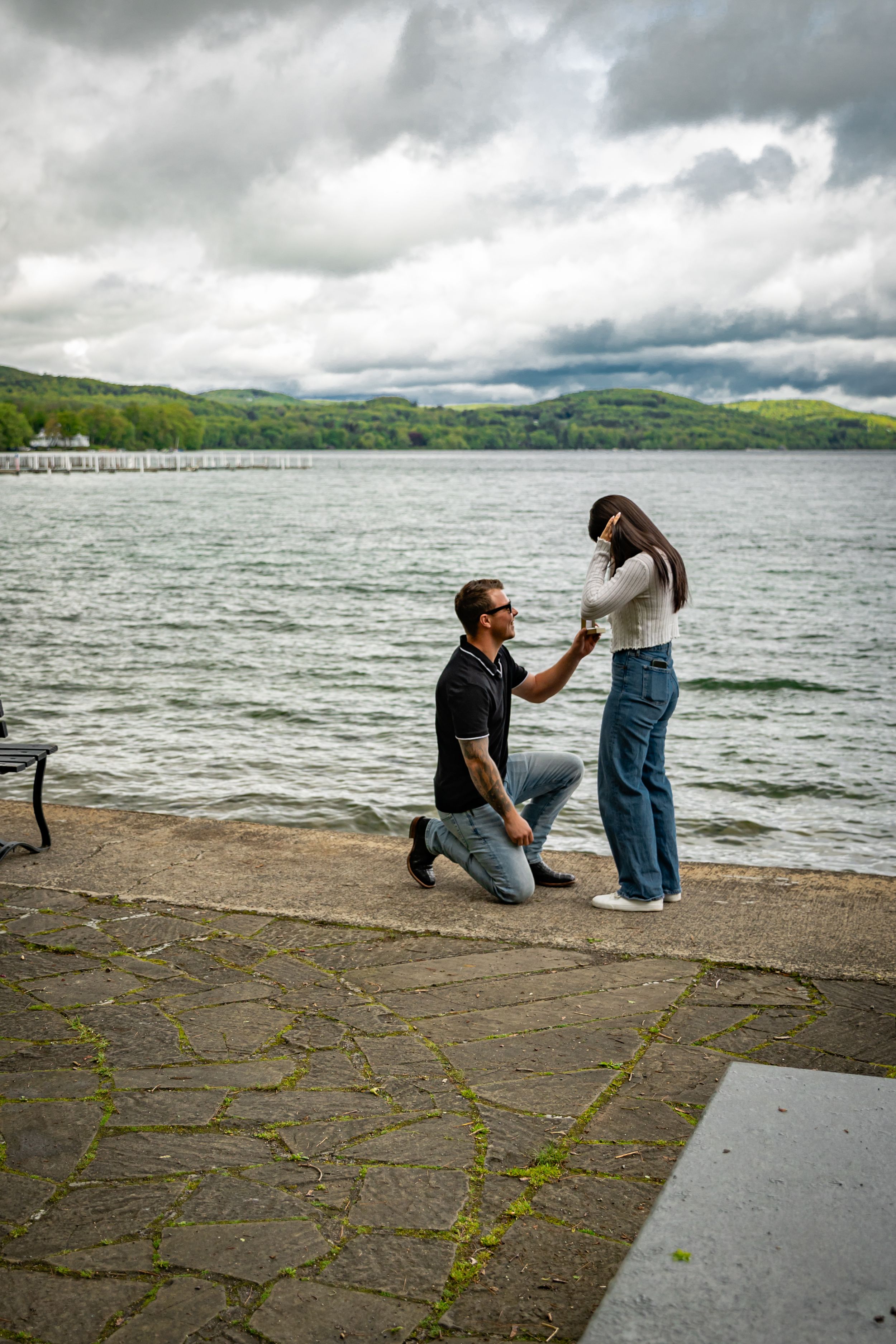 Man proposing on one knee to his girlfriend by a scenic lakeside on a cloudy day.
