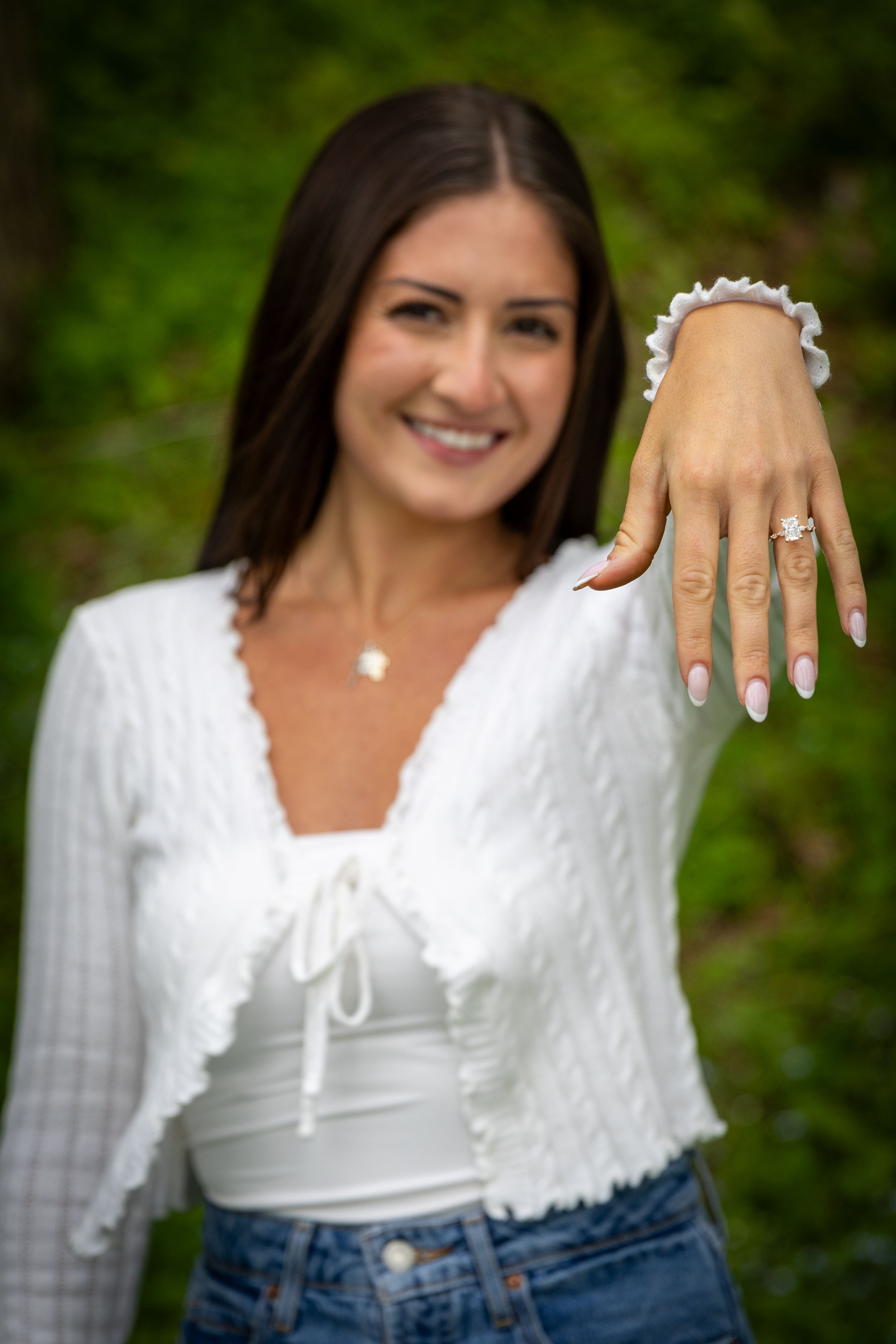 Smiling brunette woman proudly displaying her diamond engagement ring toward the camera outdoors.