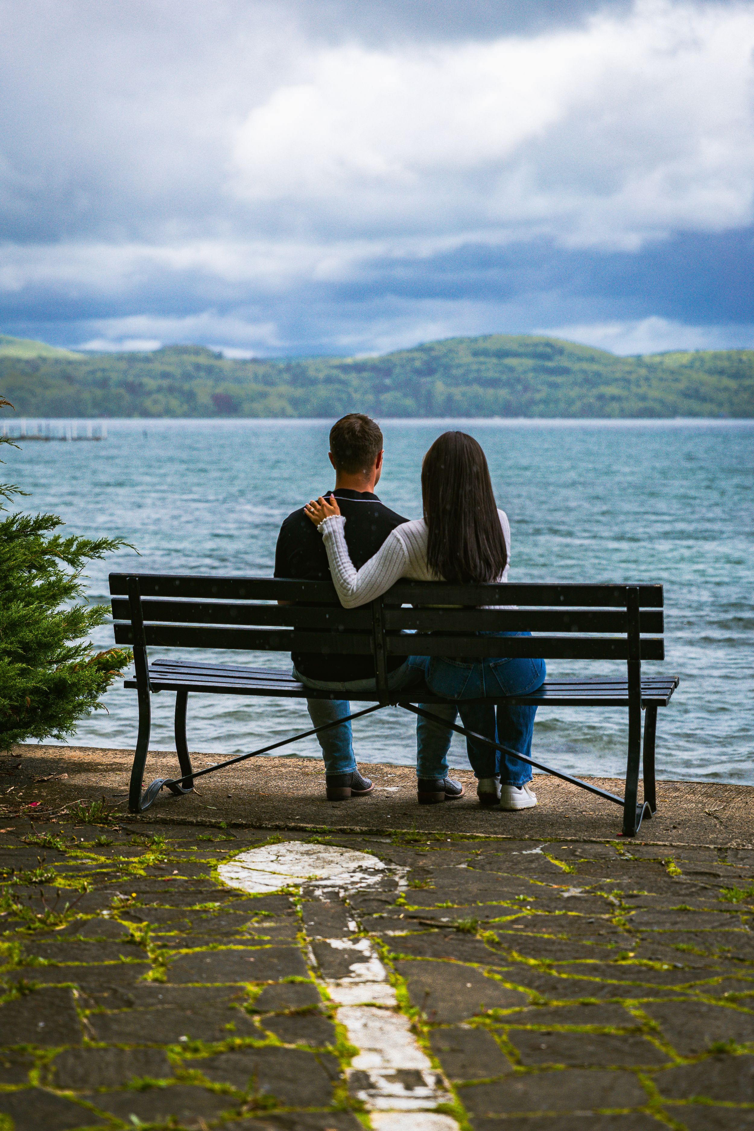 Couple sitting on bench from behind, embracing while overlooking scenic lake and green hills.