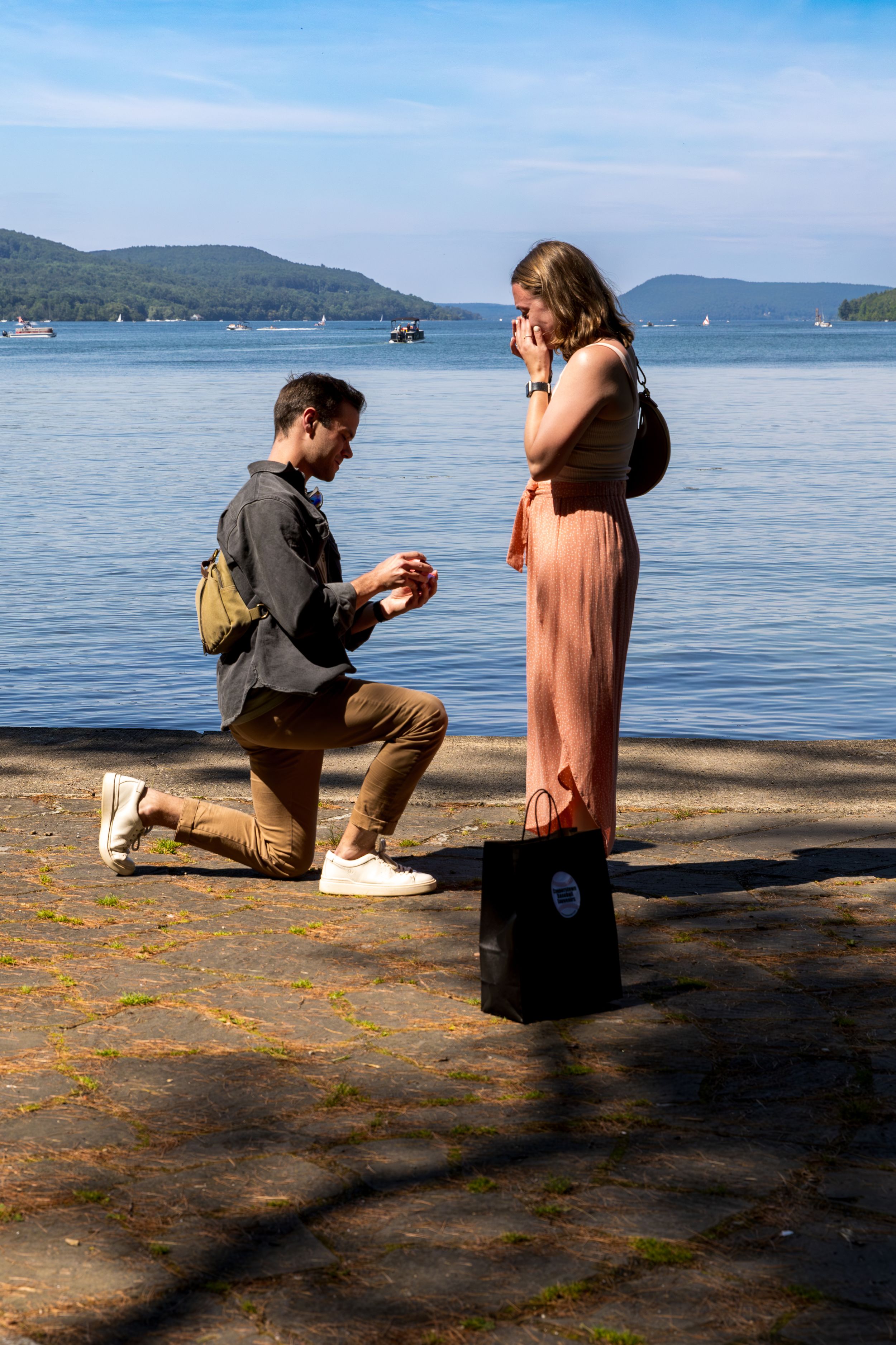 Man kneeling on one knee proposing to woman in pink dress by a scenic lakeside.