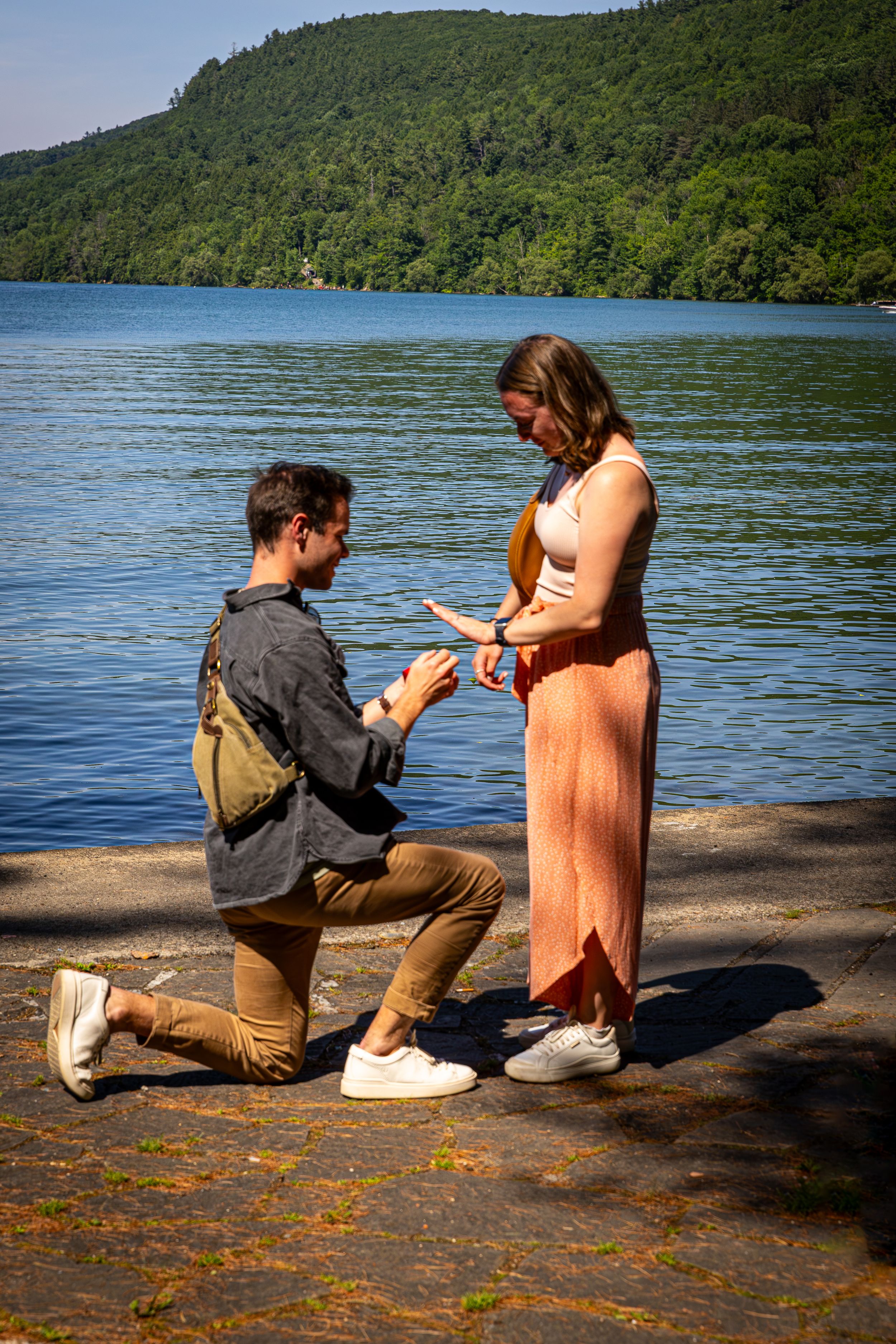 Man kneeling to place engagement ring on womans finger beside a sunlit blue lake.