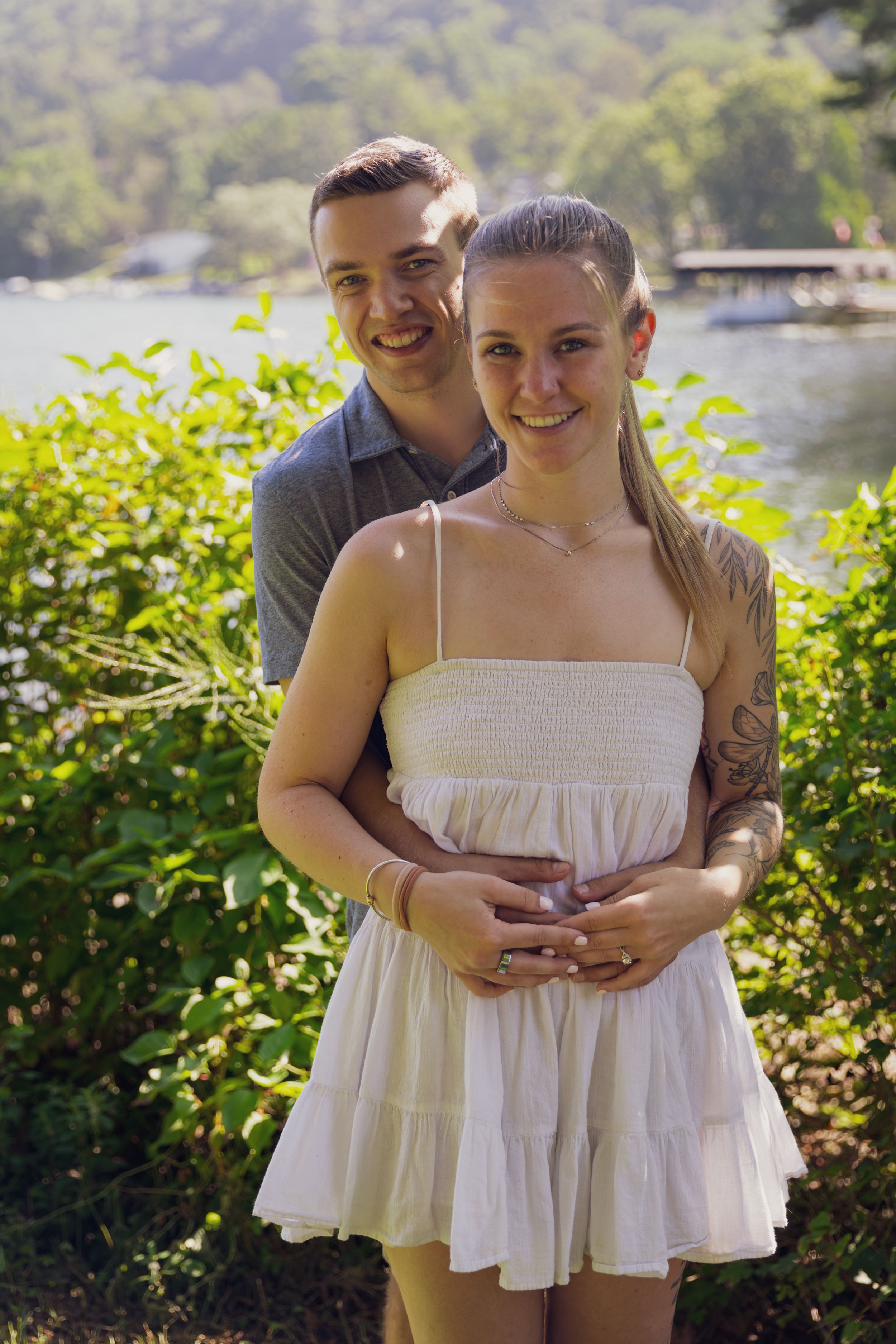 A smiling couple poses together outdoors near a lake, the man embracing the woman from behind among green foliage.