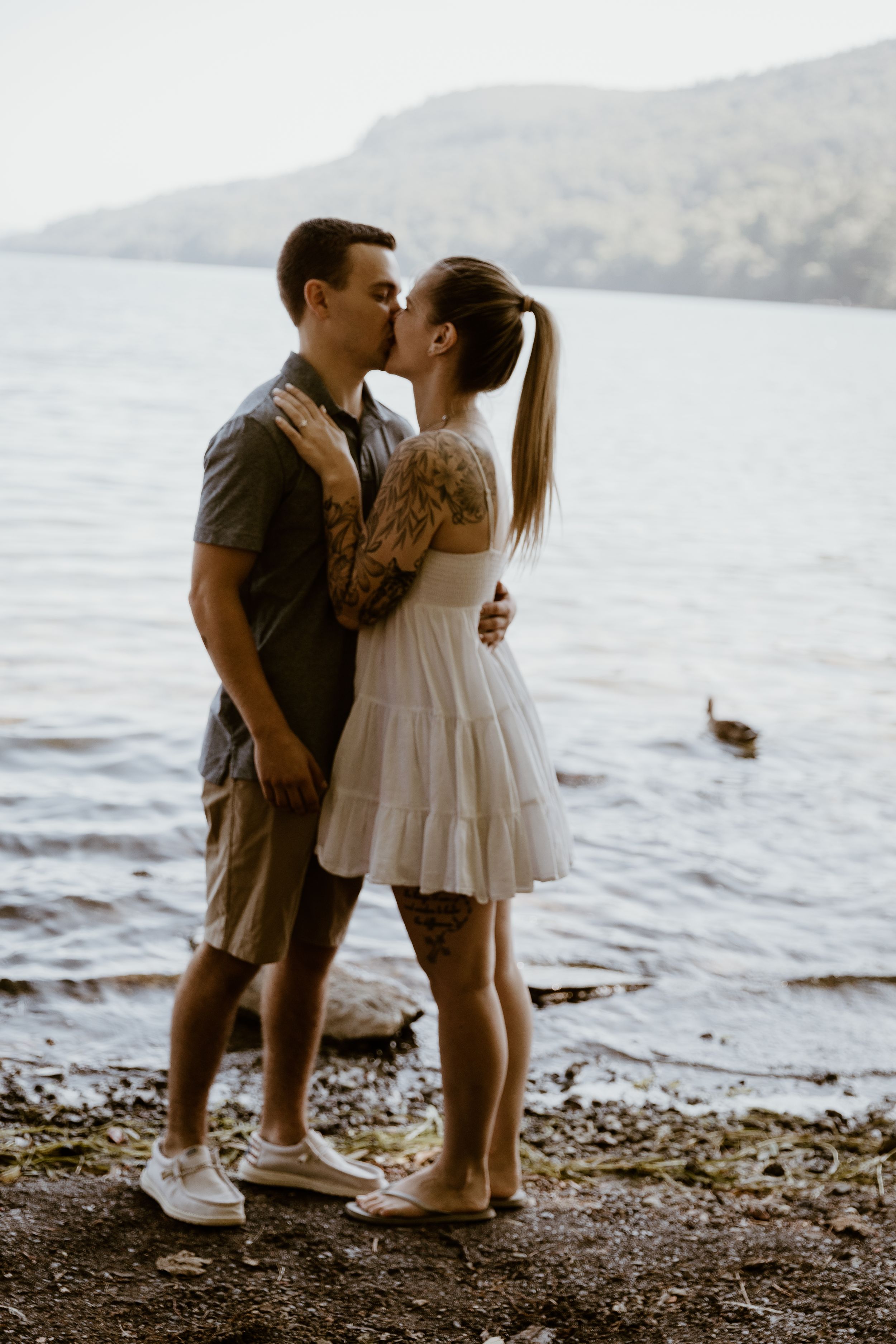 A couple shares a romantic kiss while standing on a rocky lakeshore with misty mountains visible in the background.