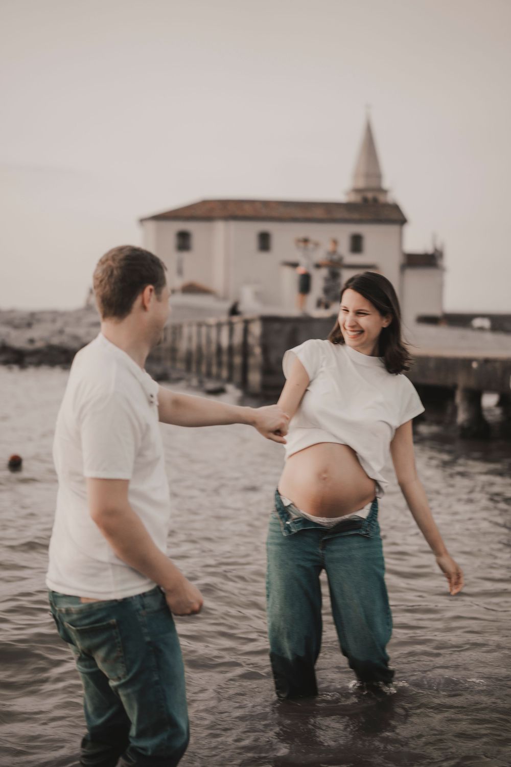Alt Text: Cinematic perspective of an elegant couple walking down the shoreline with the historic Caorle sea church.