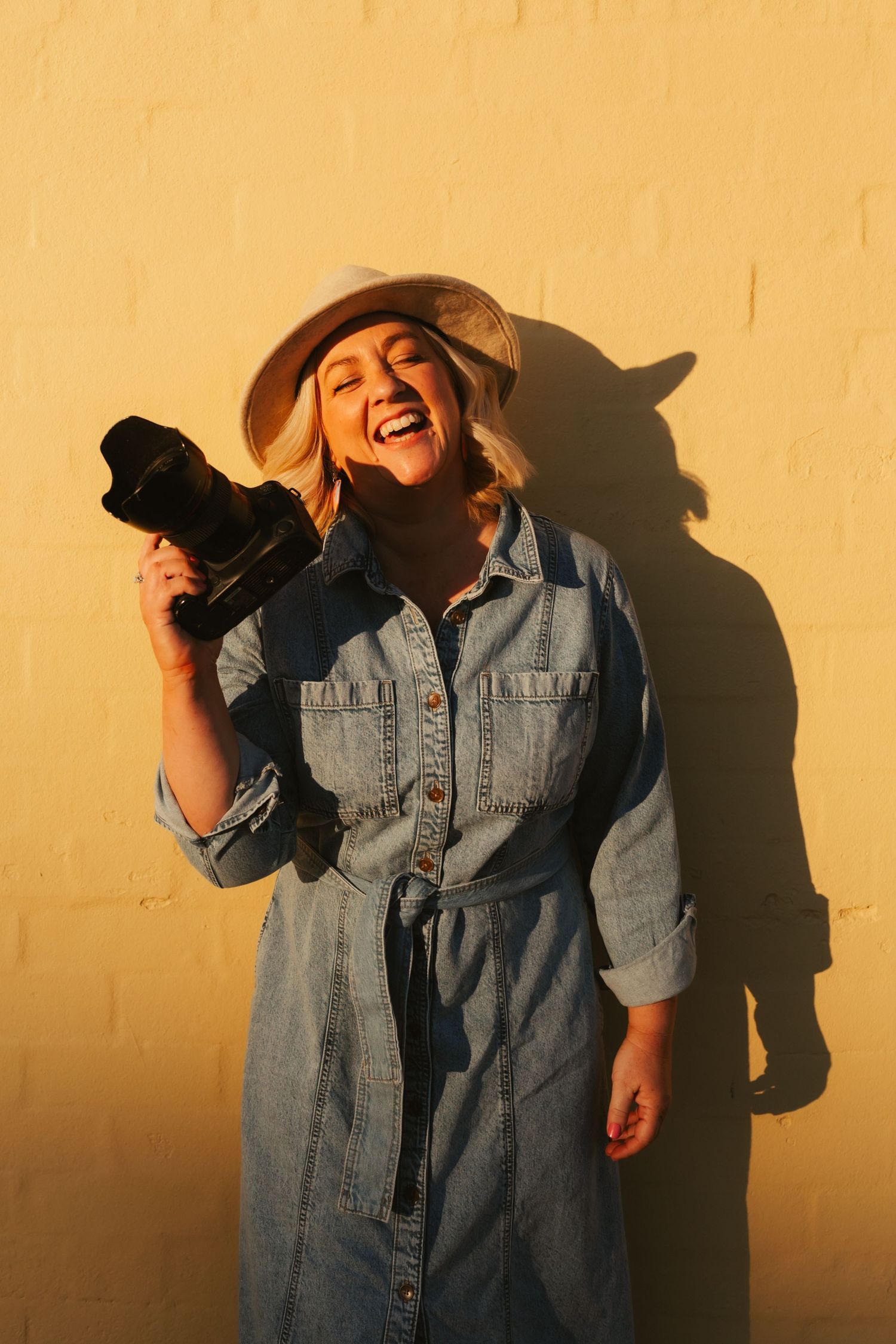 A photographer in a white shirt and sun hat laughs while looking at their camera in a golden field.