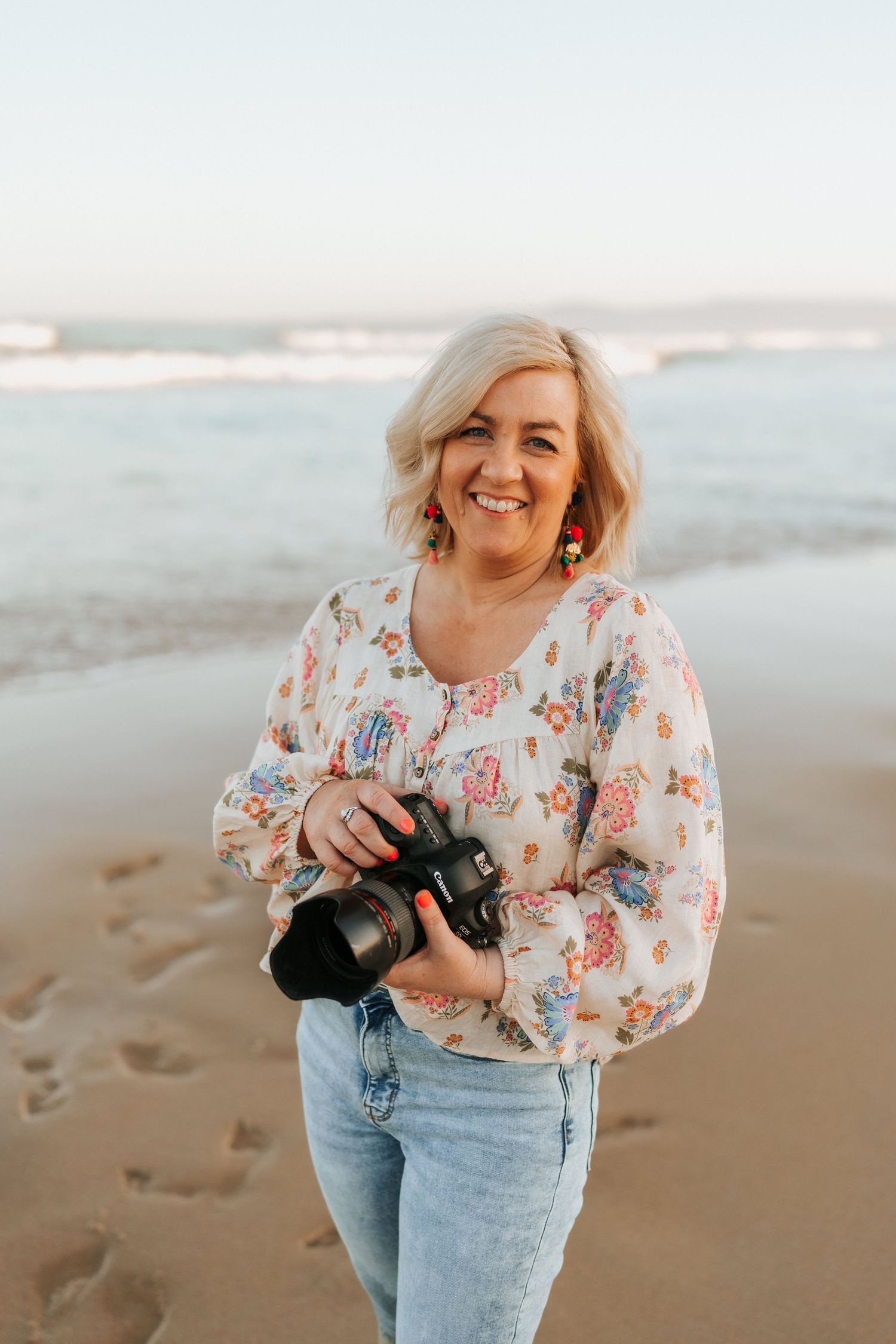 A photographer in a white shirt and sun hat laughs while looking at their camera in a golden field.