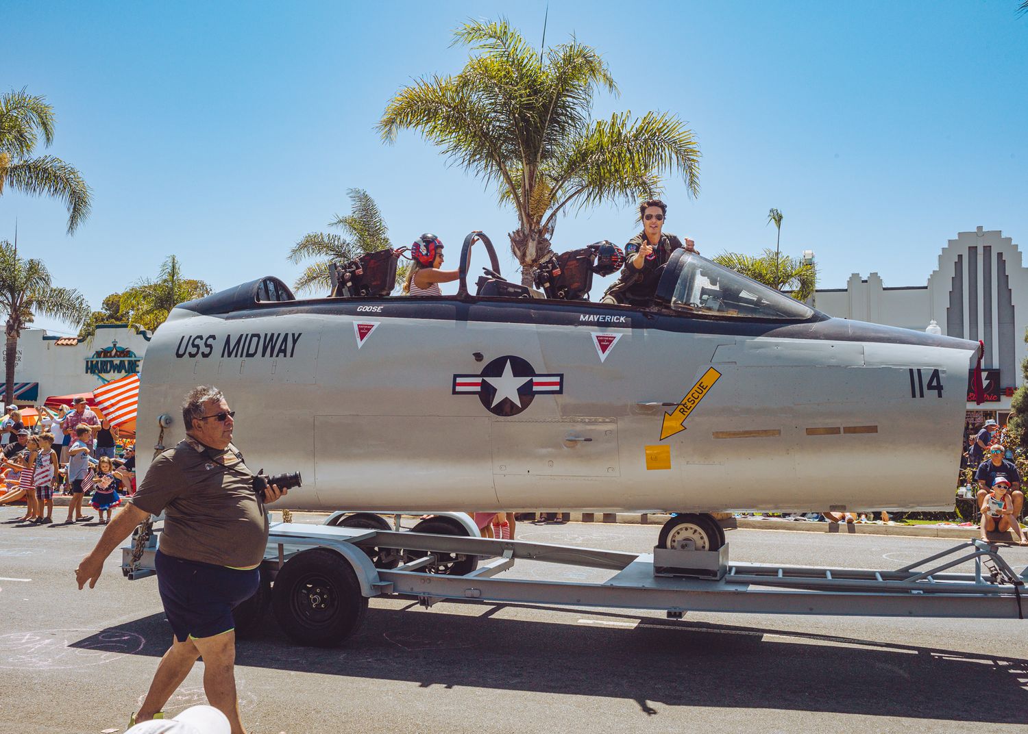 Military aircraft and palm trees line up along an outdoor display area at an airshow event.