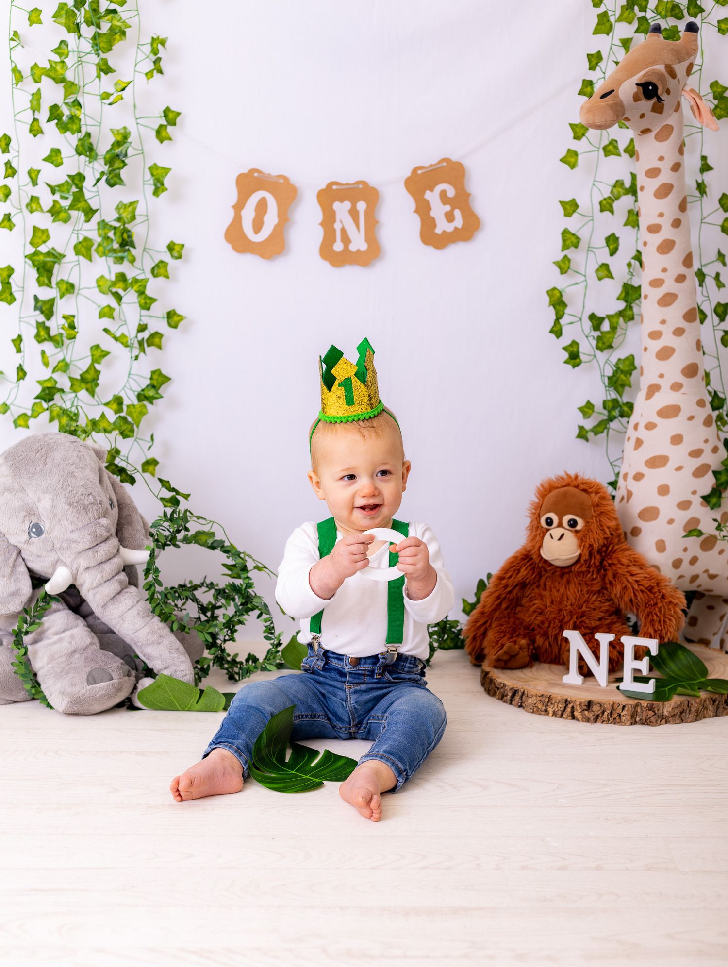 Baby's first birthday photo session setup with safari theme decorations, greenery, and plush animal toys.