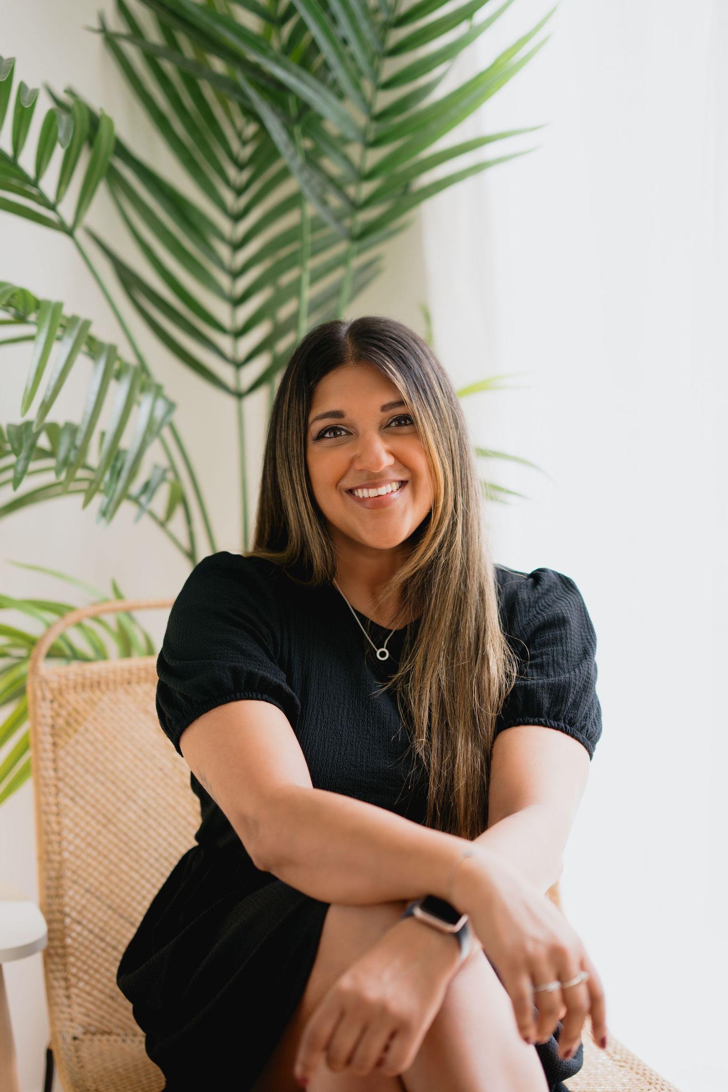 A person in a black top sits in a rattan chair against a white wall with tropical palm leaves in the background.