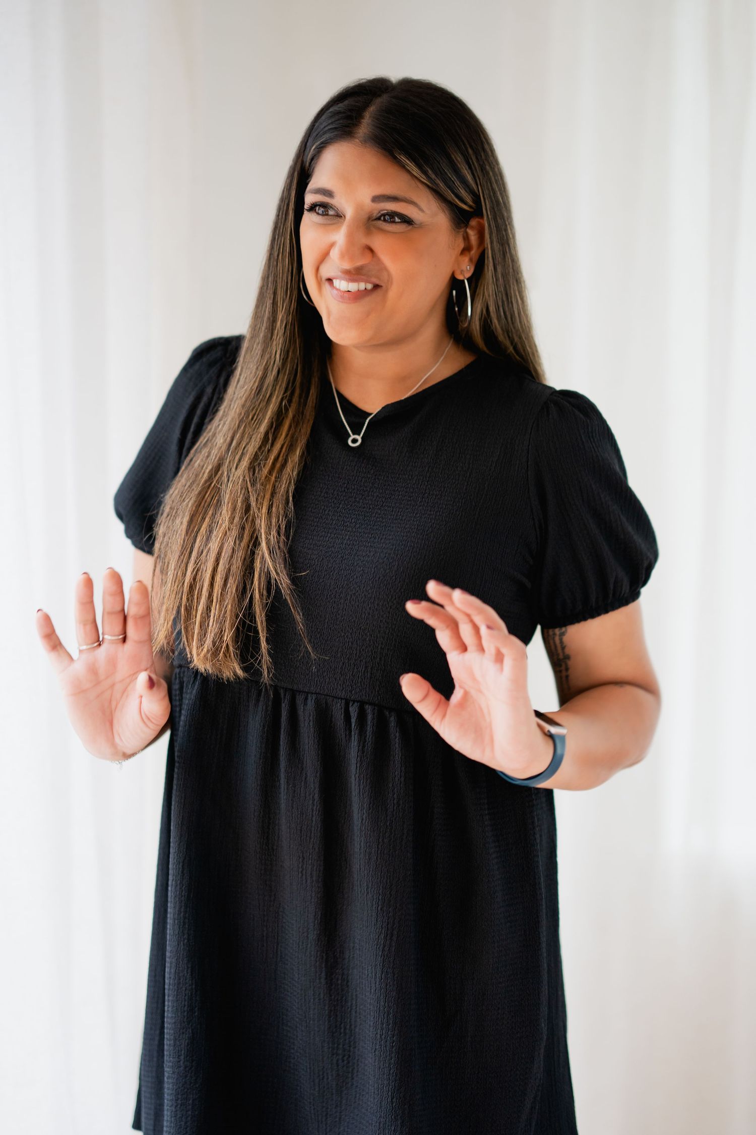A person wearing a black dress gestures playfully with raised hands against a white background.