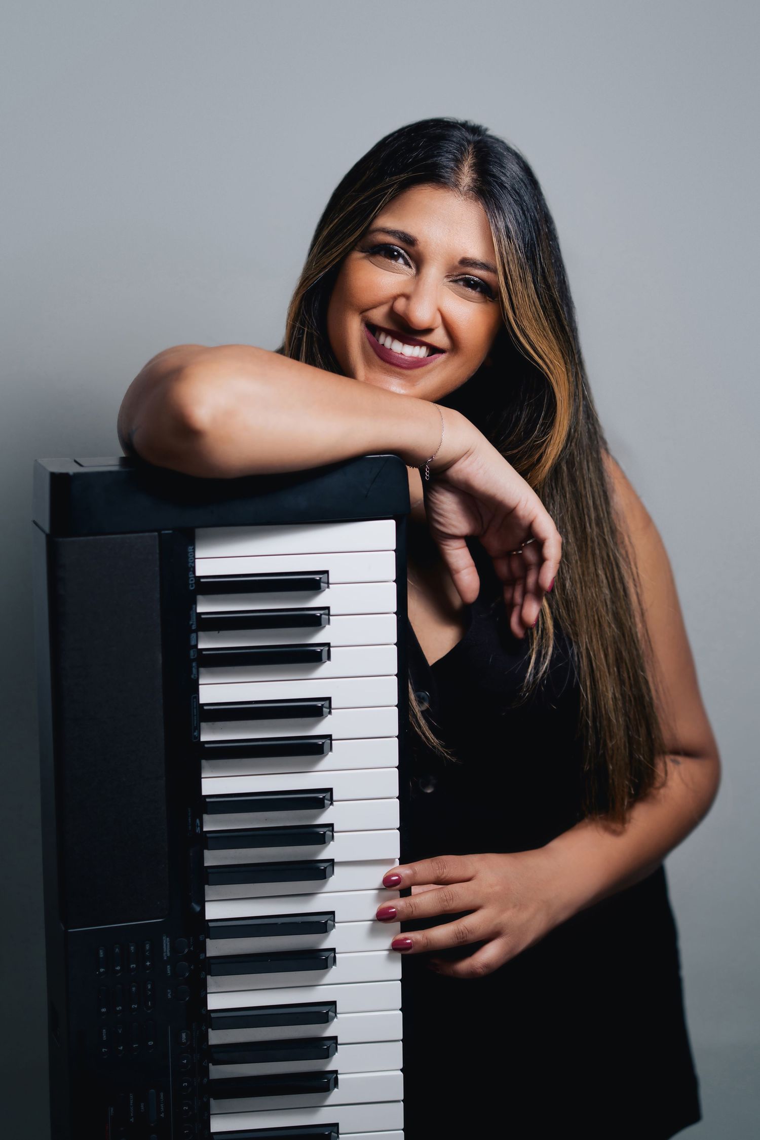 Musician in black top poses joyfully with digital piano keyboard against gray studio background.