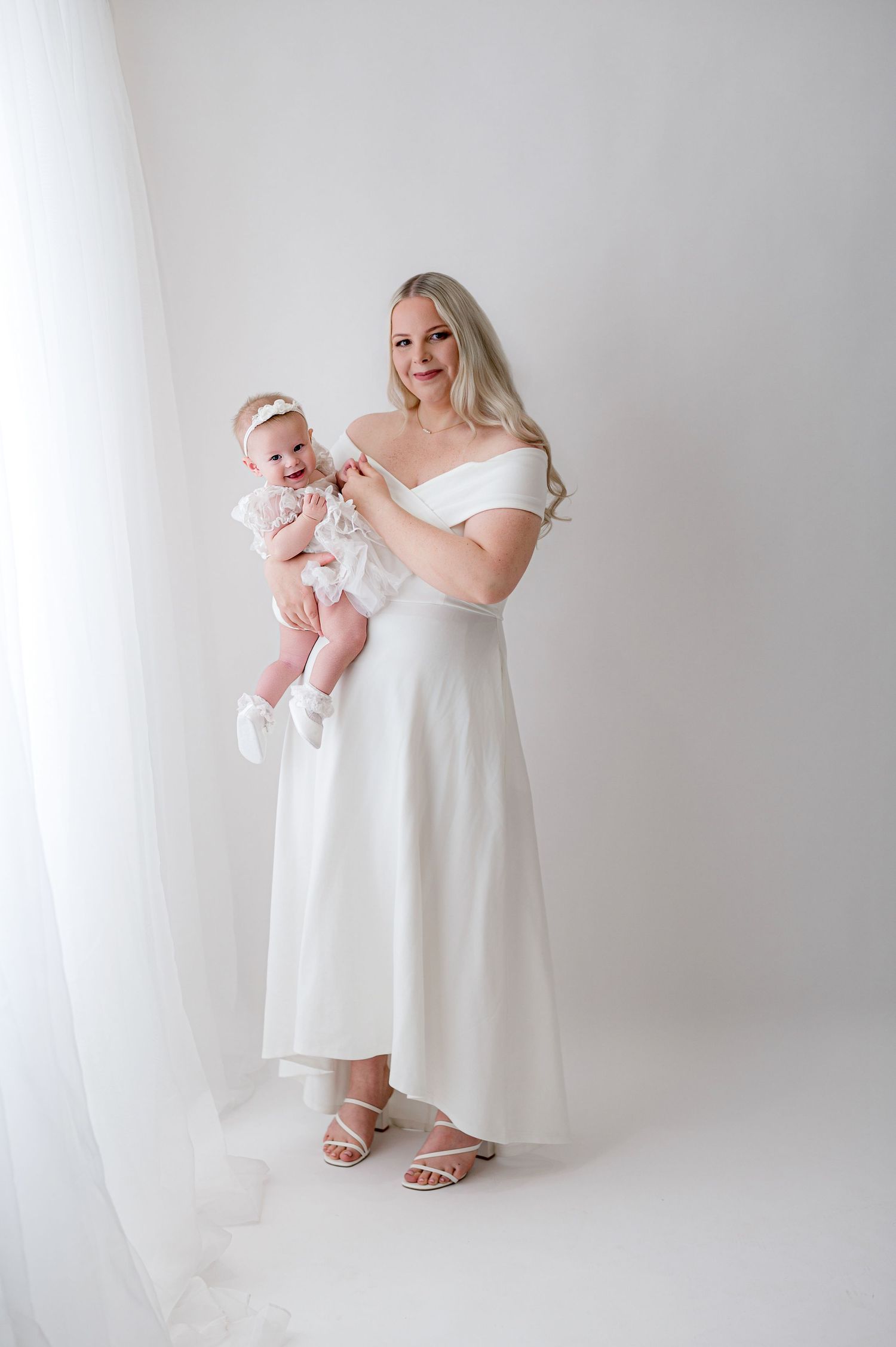 A person in a white lace dress sits on a white chair holding a baby in a minimalist studio setting.