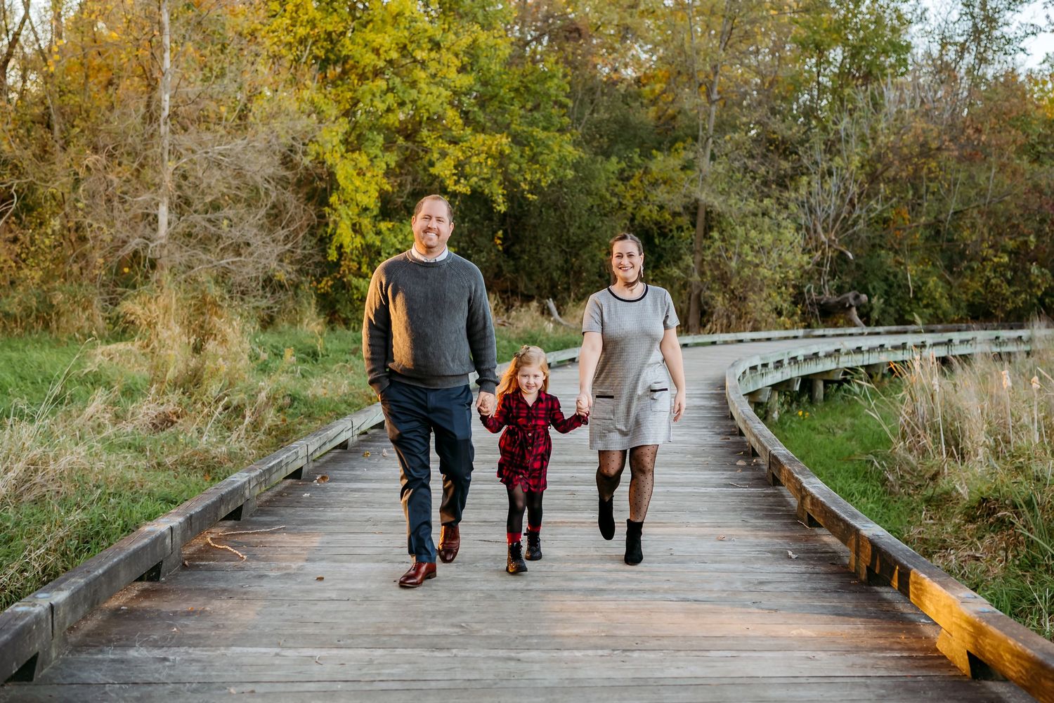A family walks together on a wooden boardwalk path surrounded by autumn foliage and trees.