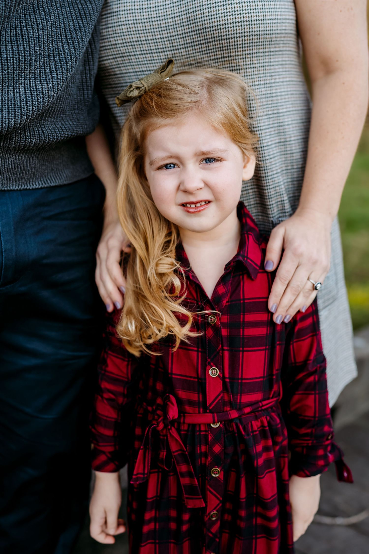 A child wearing a red and black buffalo plaid dress stands next to an adult in casual clothing outdoors.