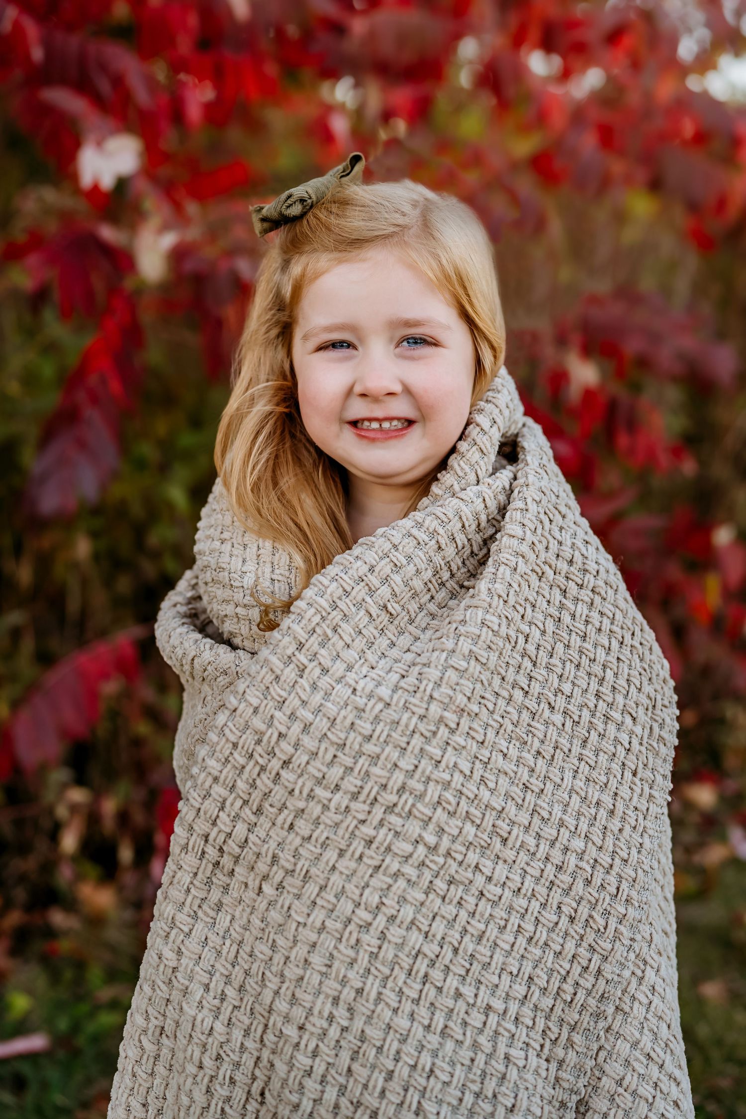 Child wrapped in cozy beige blanket smiles against backdrop of bright red autumn leaves.