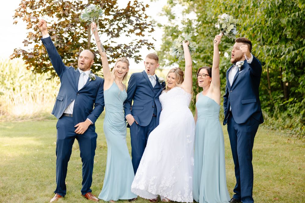 Wedding party in light blue bridesmaid dresses and navy suits pose together outdoors on a sunny day.