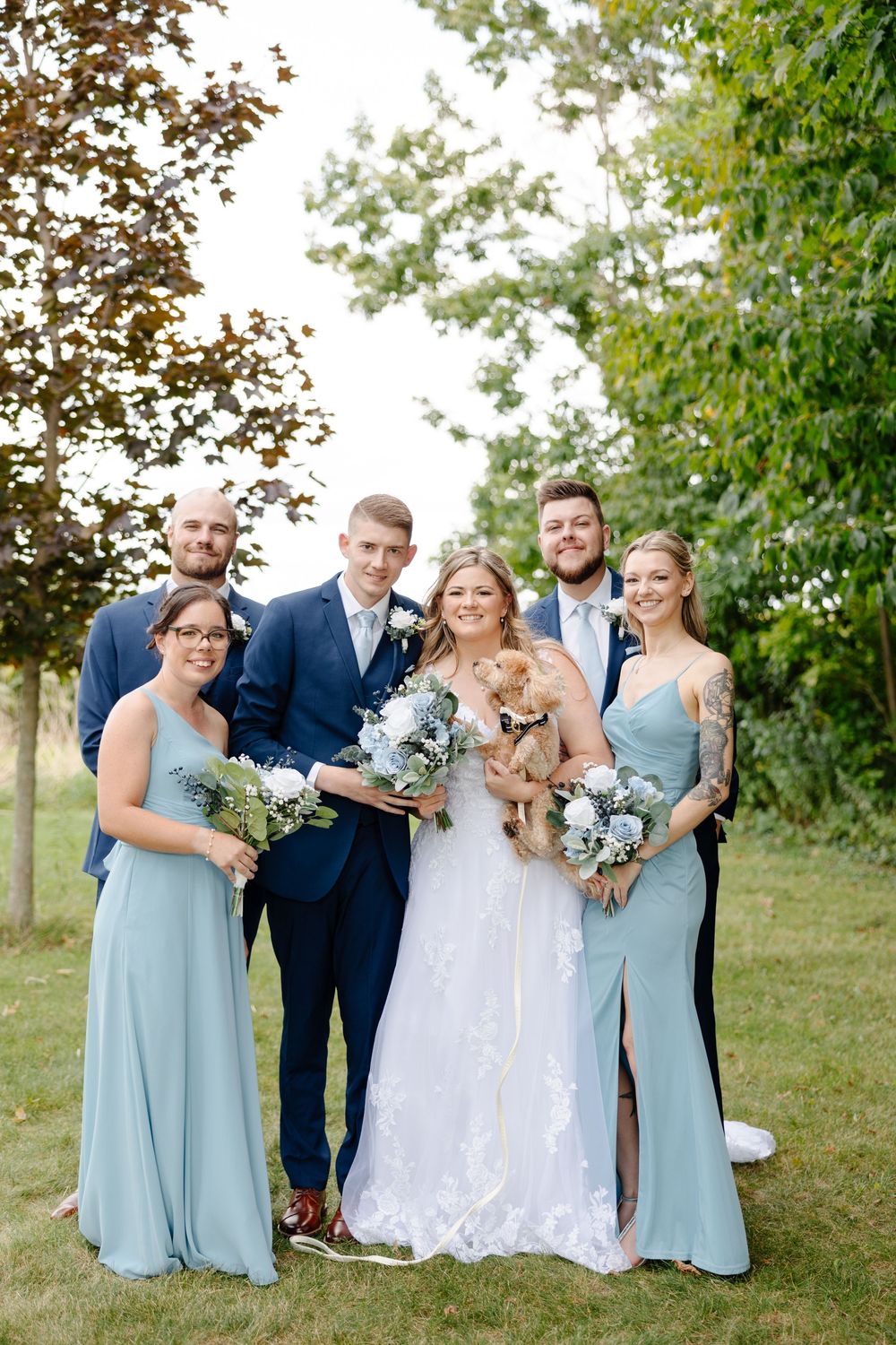 Wedding group stands together on grassy hill with bridesmaids in light blue dresses holding bouquets.
