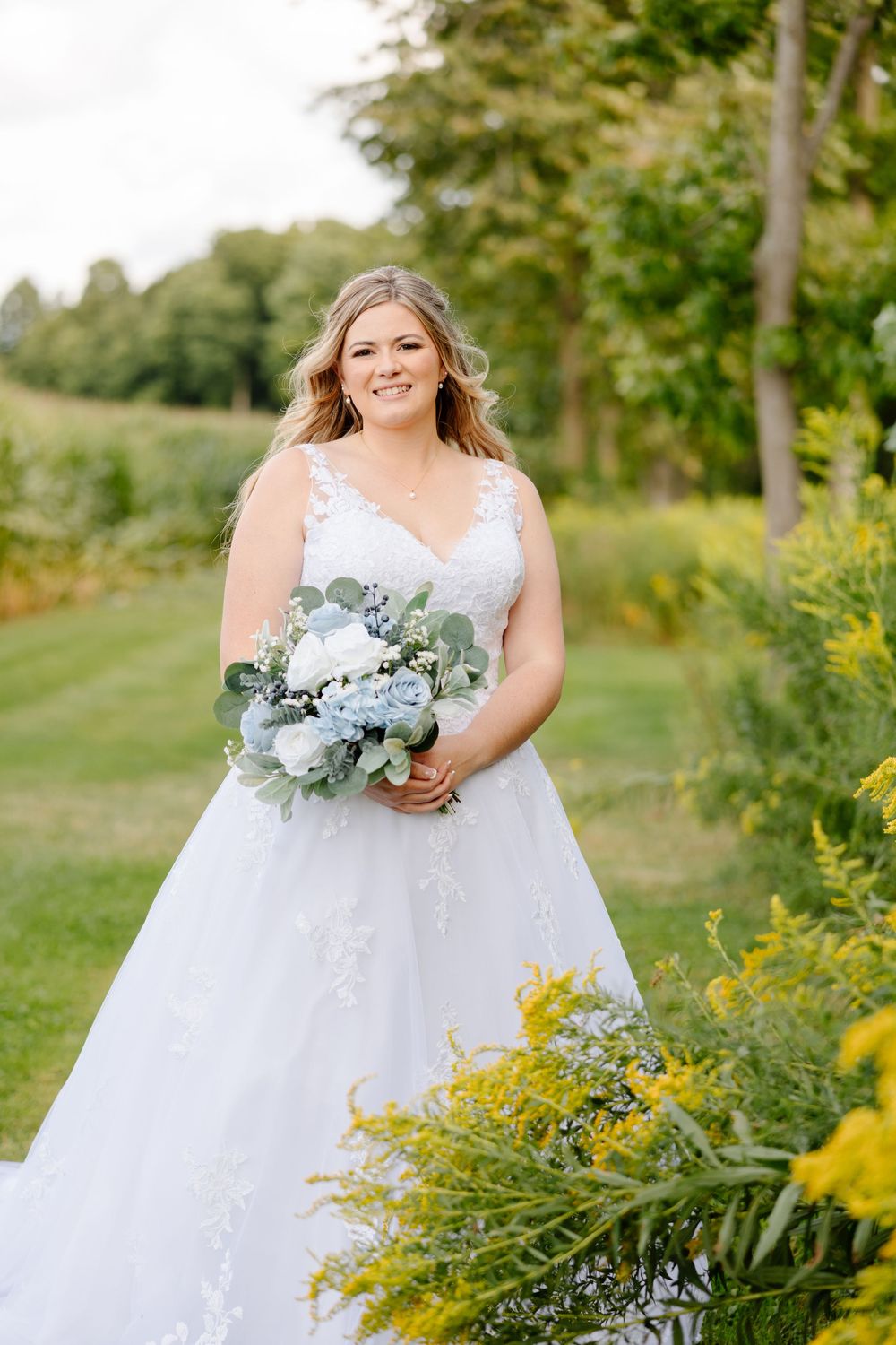 A bride in a flowing white gown poses in a garden setting with blue and green floral bouquet.