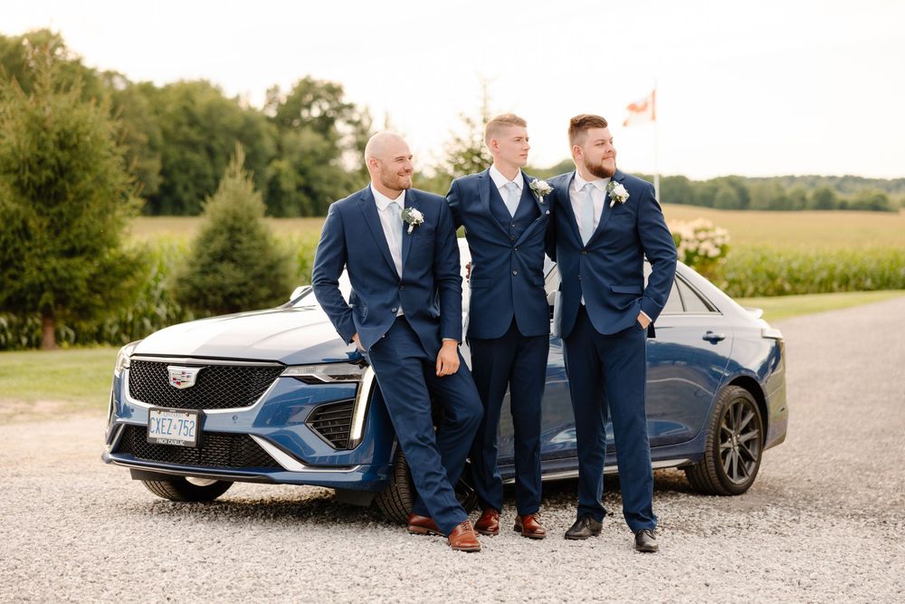 Groomsmen in navy suits pose with luxury car on rural road at sunset.