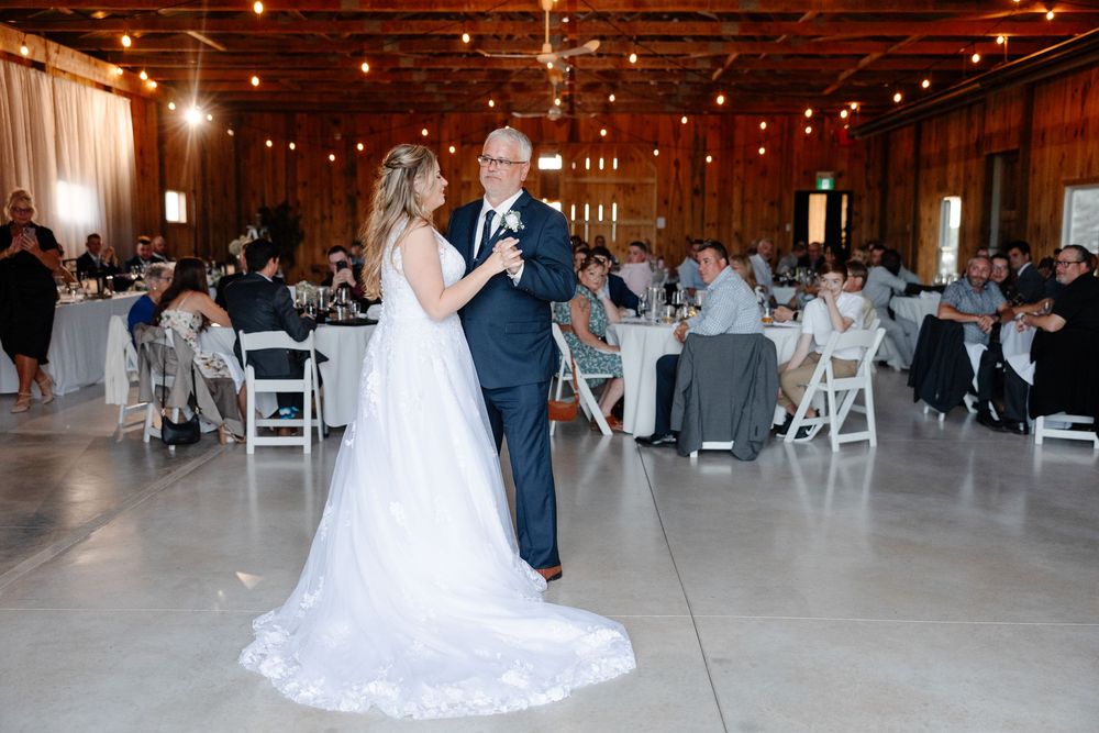 A couple shares their first dance at a rustic barn wedding reception with string lights overhead and white chairs.