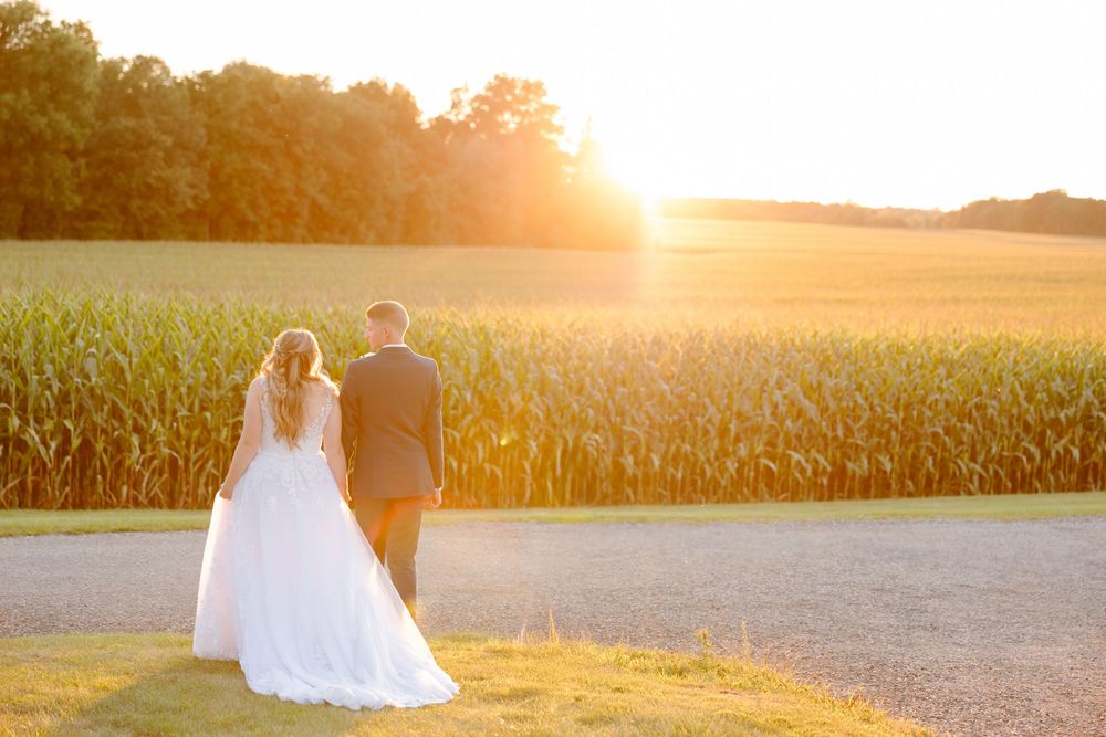 A couple in wedding attire stands silhouetted against a golden sunset in a rural field during their special day.
