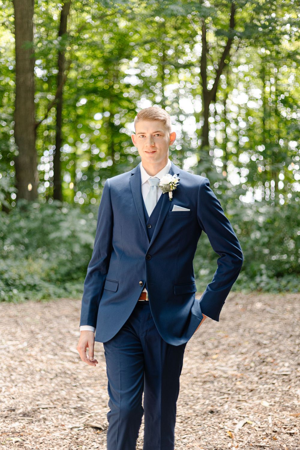Two people in navy blue suits stand in a wooded outdoor setting with sunlit trees in the background.