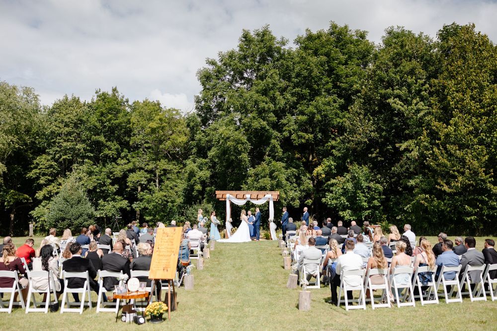 Panoramic view of an outdoor wedding ceremony setup with white chairs arranged on a grassy hillside under trees.