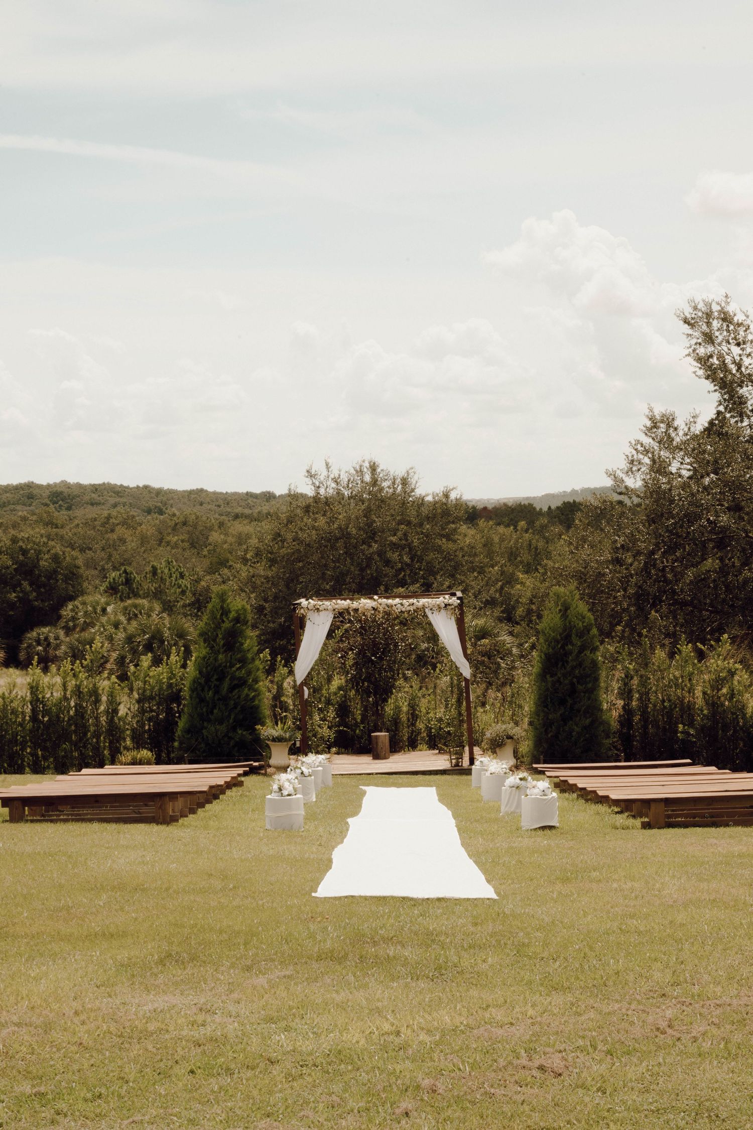 Outdoor wedding ceremony setup with wooden benches and white fabric arch in a scenic grassy field.
