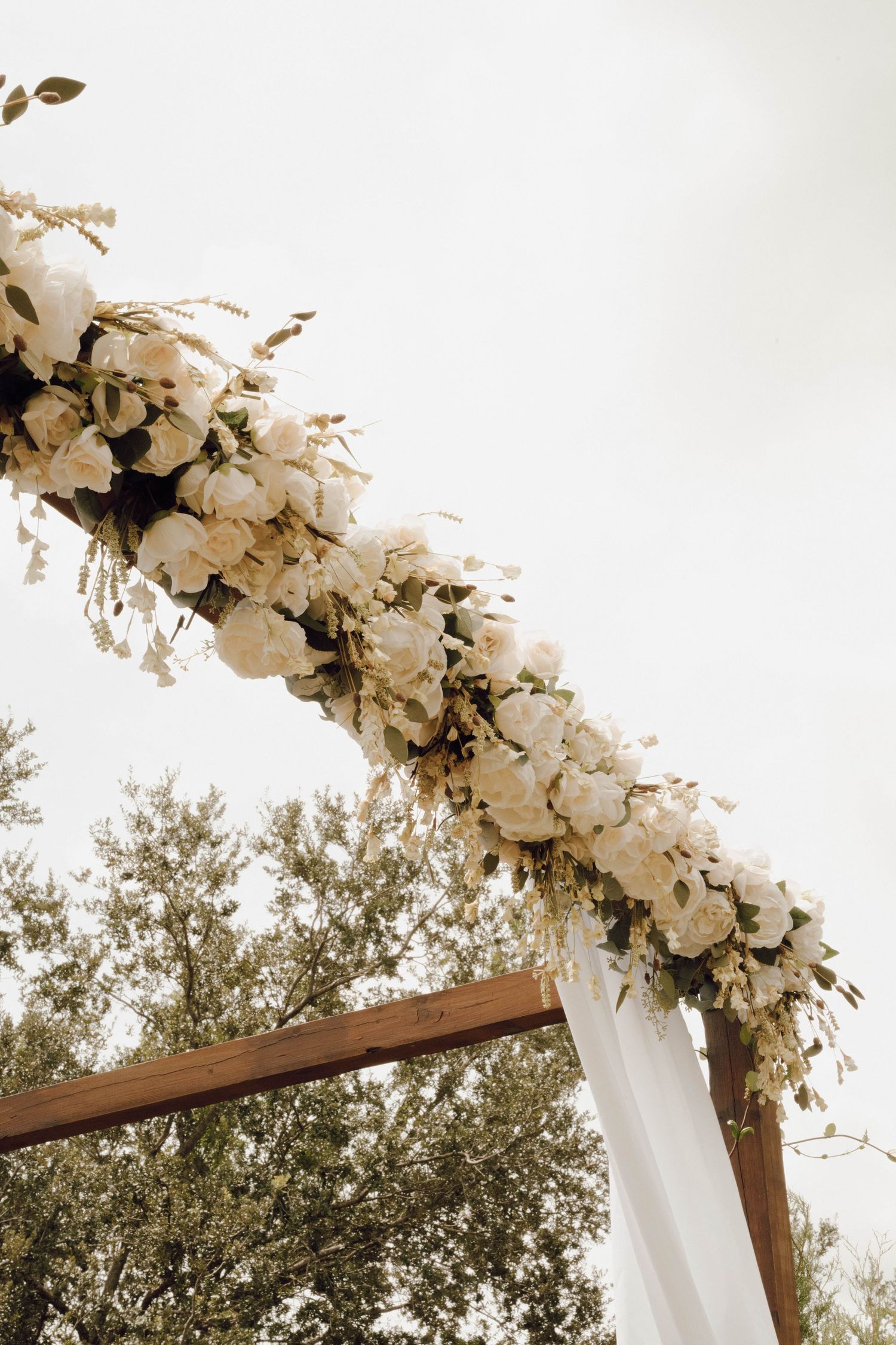 Elegant wedding arch decorated with white fabric and floral arrangements against a backdrop of trees.