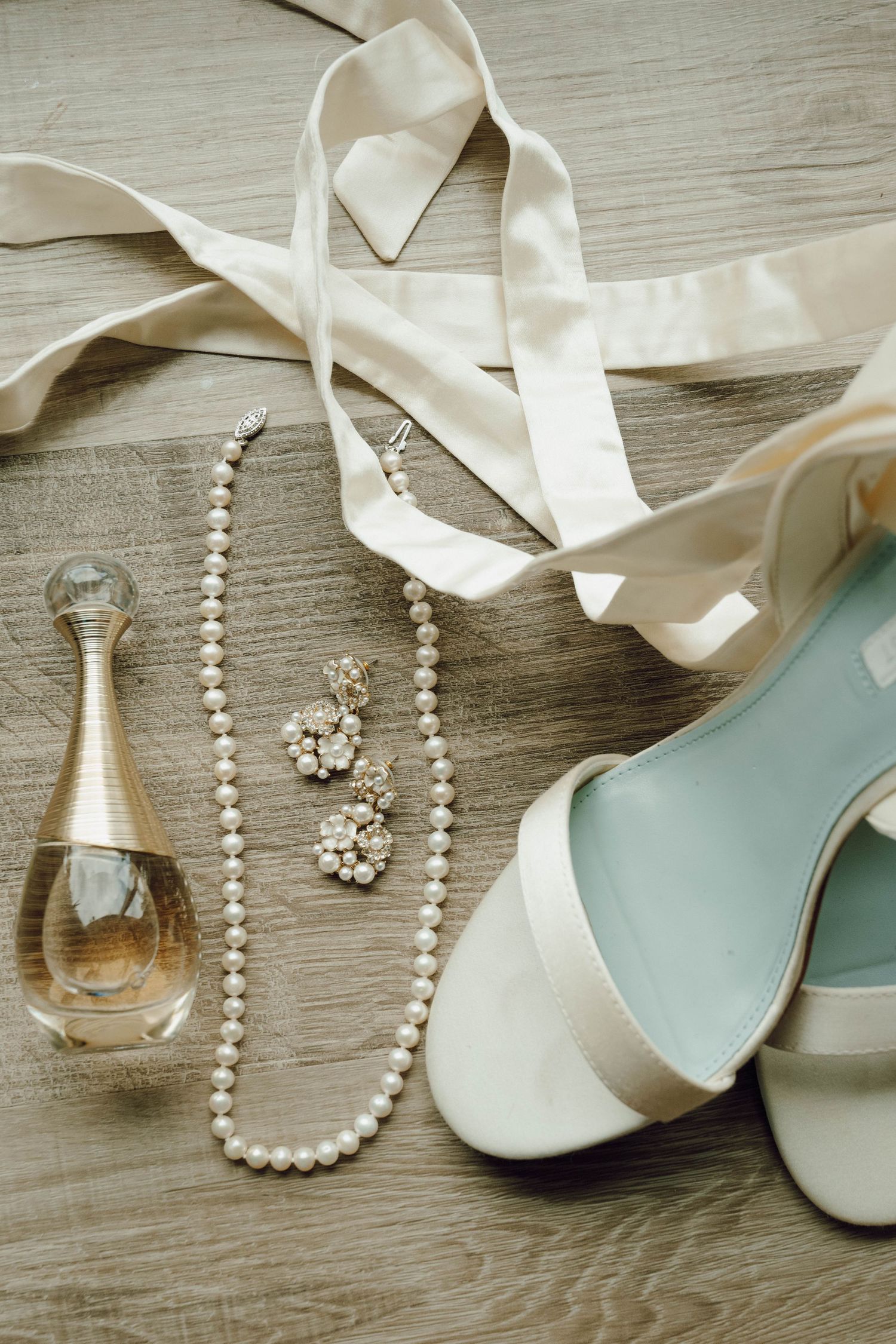 Wedding accessories including white shoes, pearl necklace, and perfume bottle arranged on a gray wooden surface.