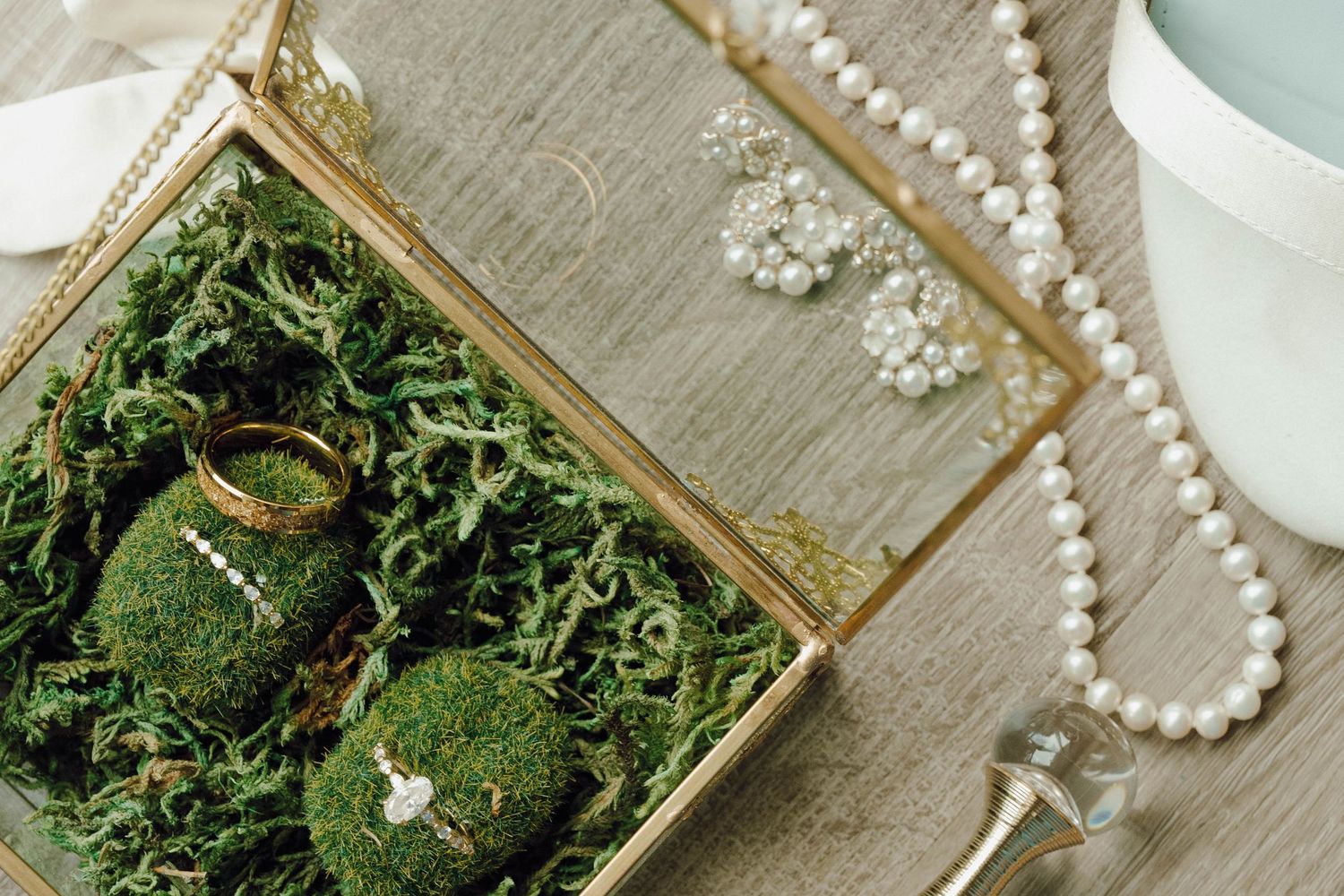 Wedding rings nestled in green moss inside an ornate gold box with pearl necklaces on wooden surface.