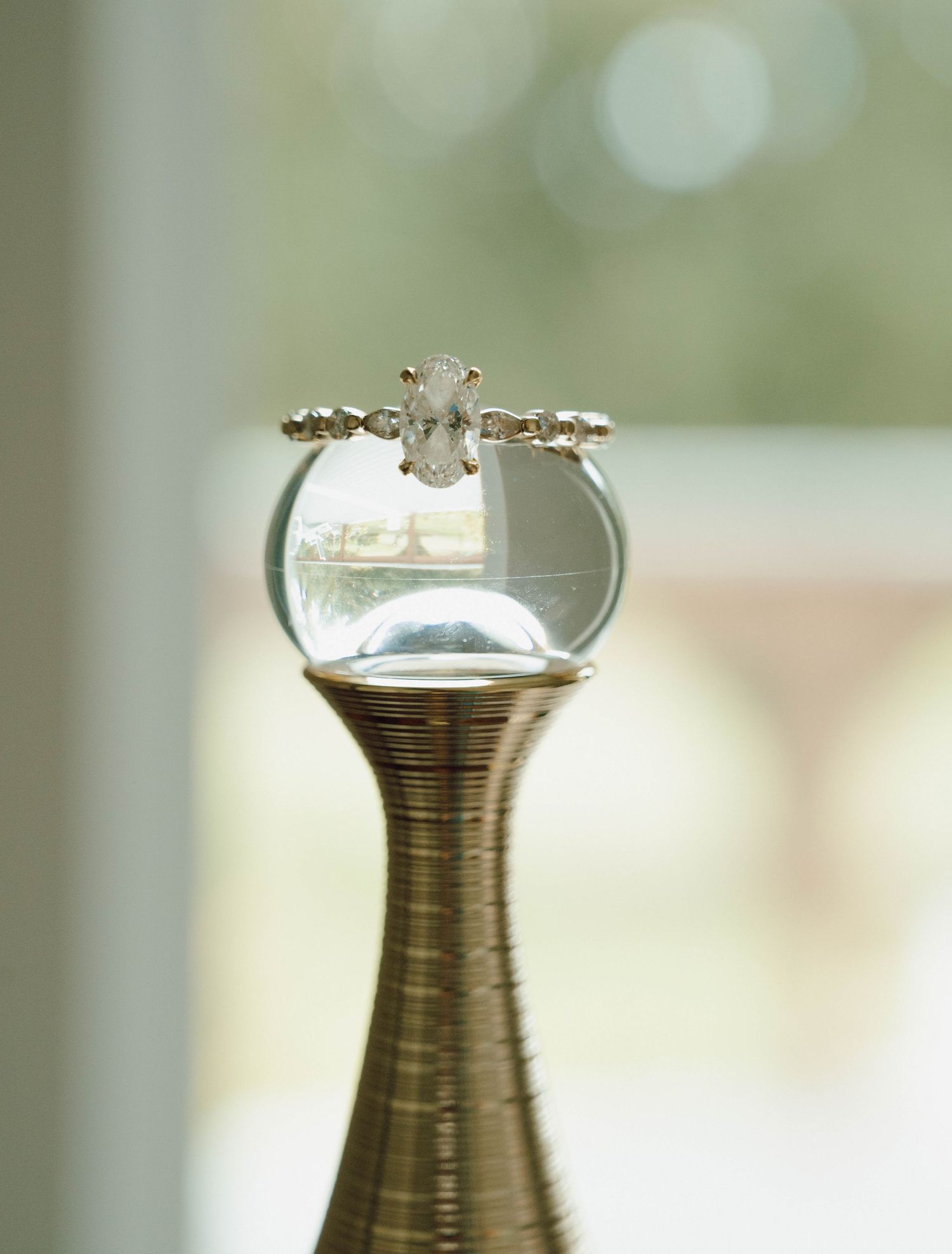 Diamond engagement ring balanced on glass vase with soft bokeh background.