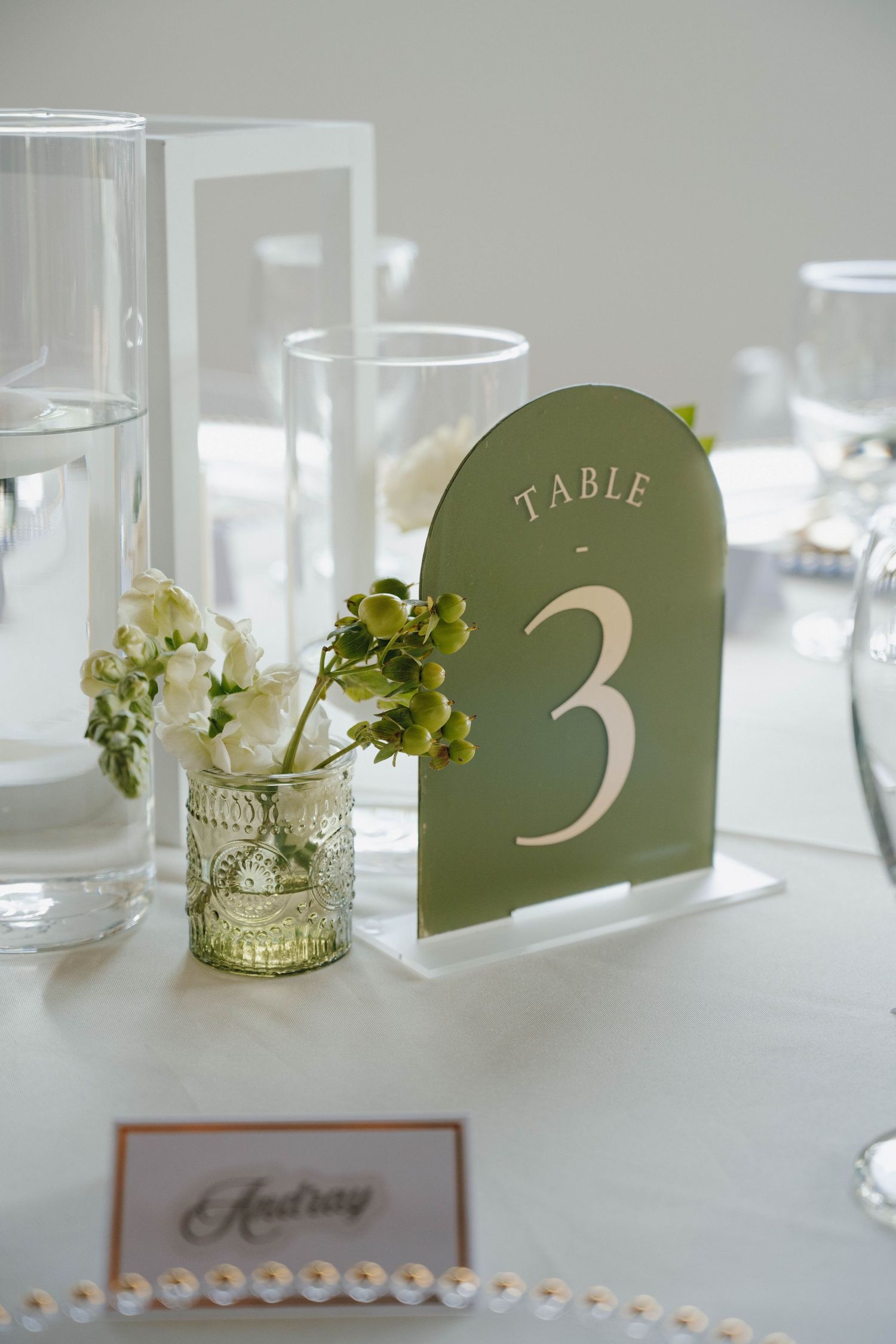 Elegant wedding table setting with green table number 3 sign and small floral arrangement in vintage glass vase.