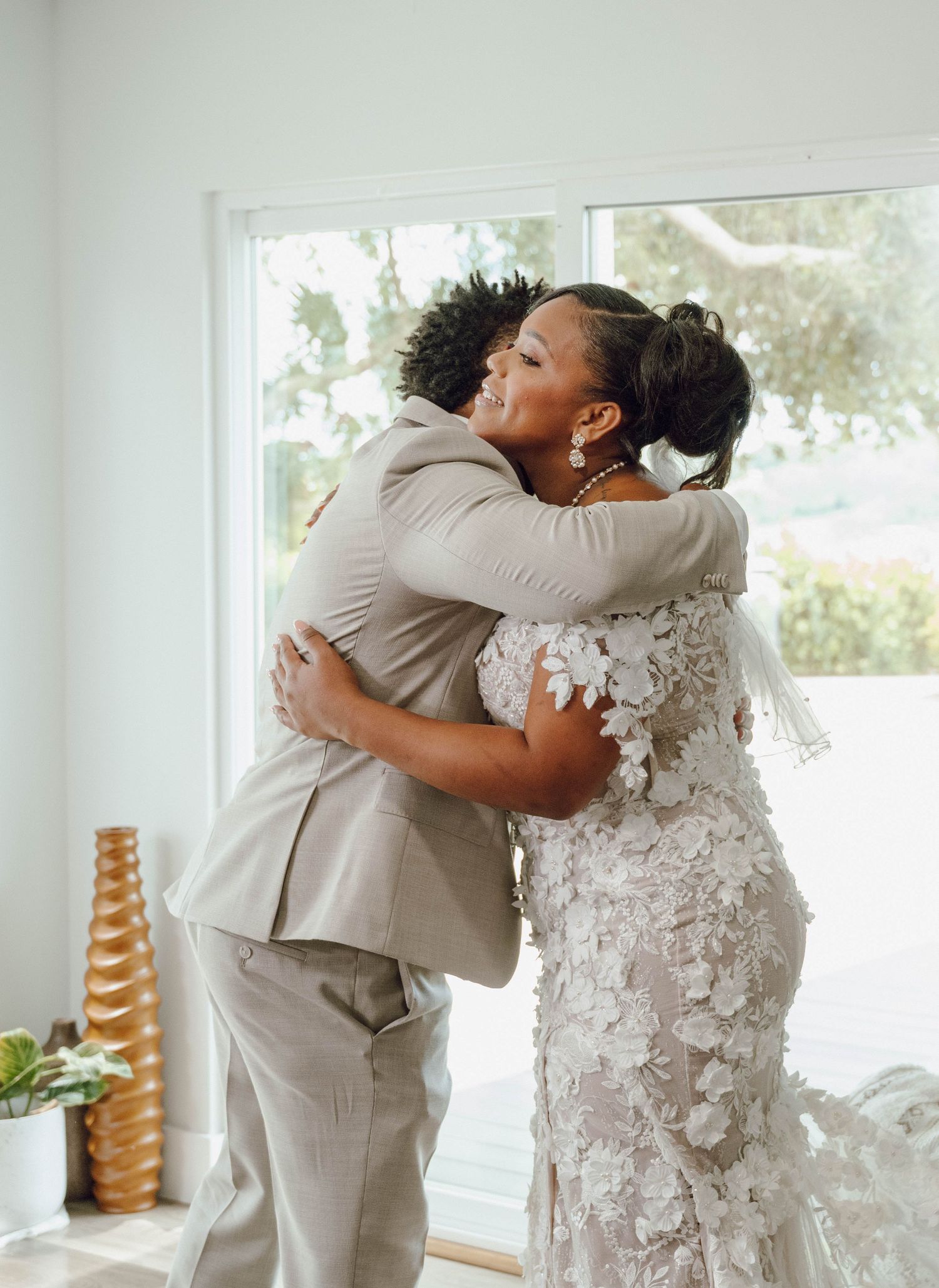 A tender embrace between two wedding guests, with one wearing a lace wedding dress and the other in a beige suit.