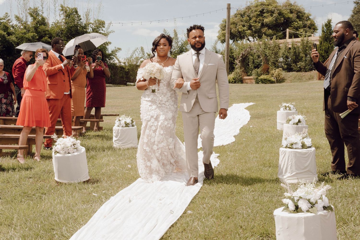 Outdoor garden wedding ceremony with white aisle runner and decorative white pedestals.