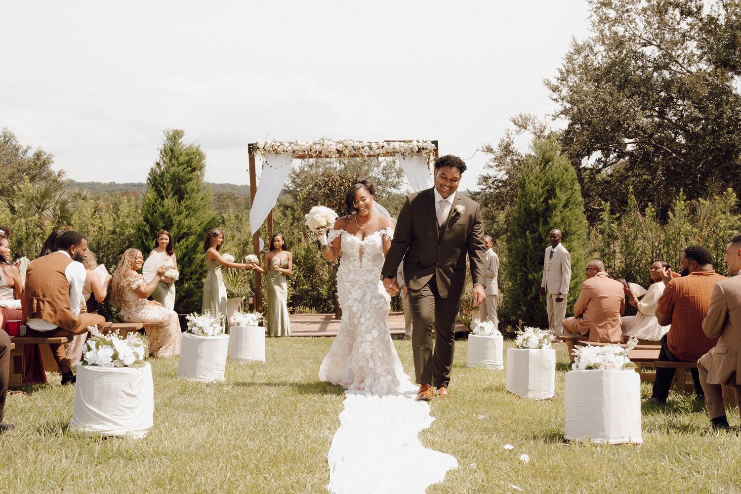 Rustic outdoor wedding ceremony setup with wooden arch and white pedestals lining the aisle.