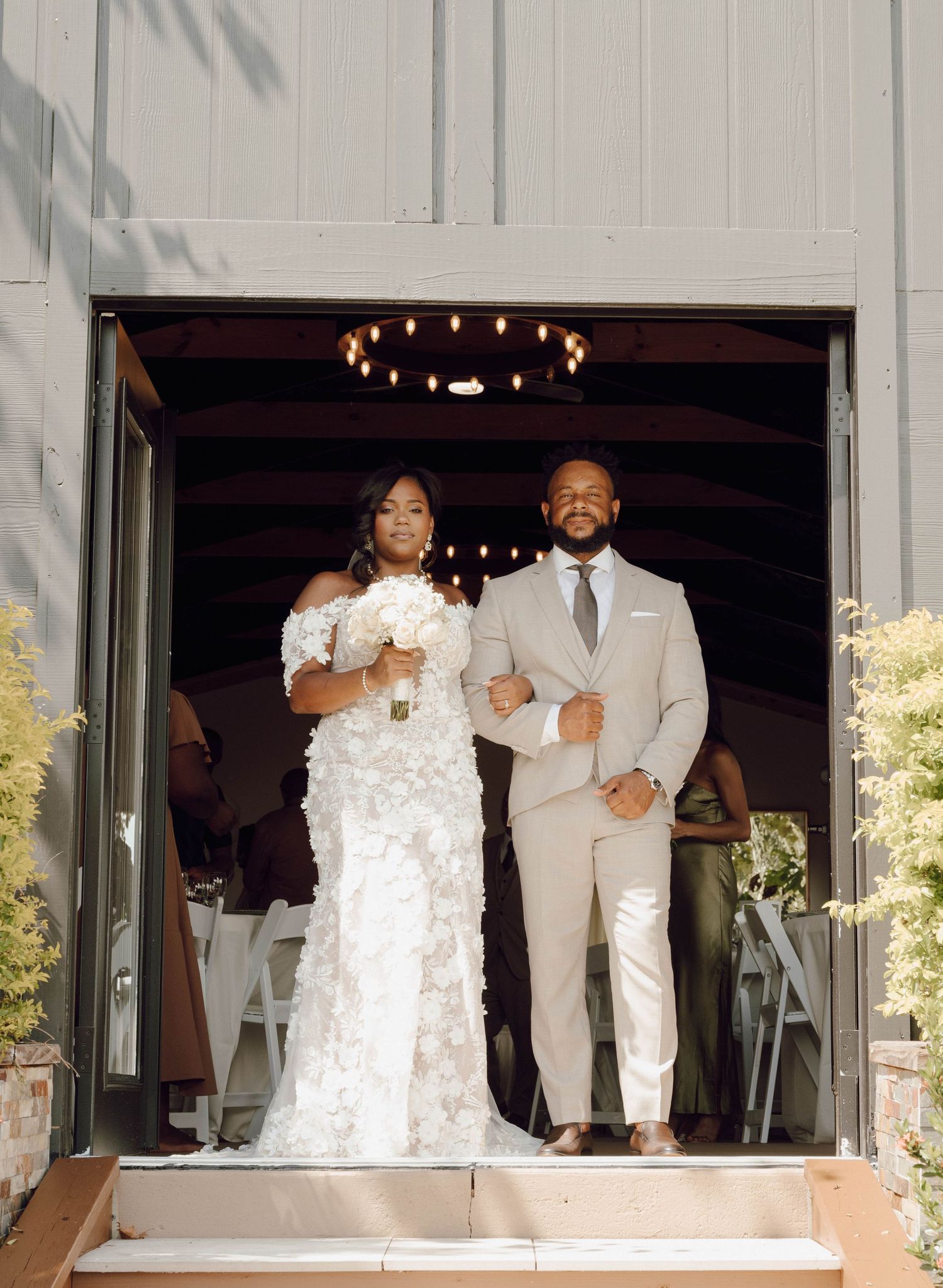 A joyful wedding couple stands together in formal white attire on a stage during their reception celebration.