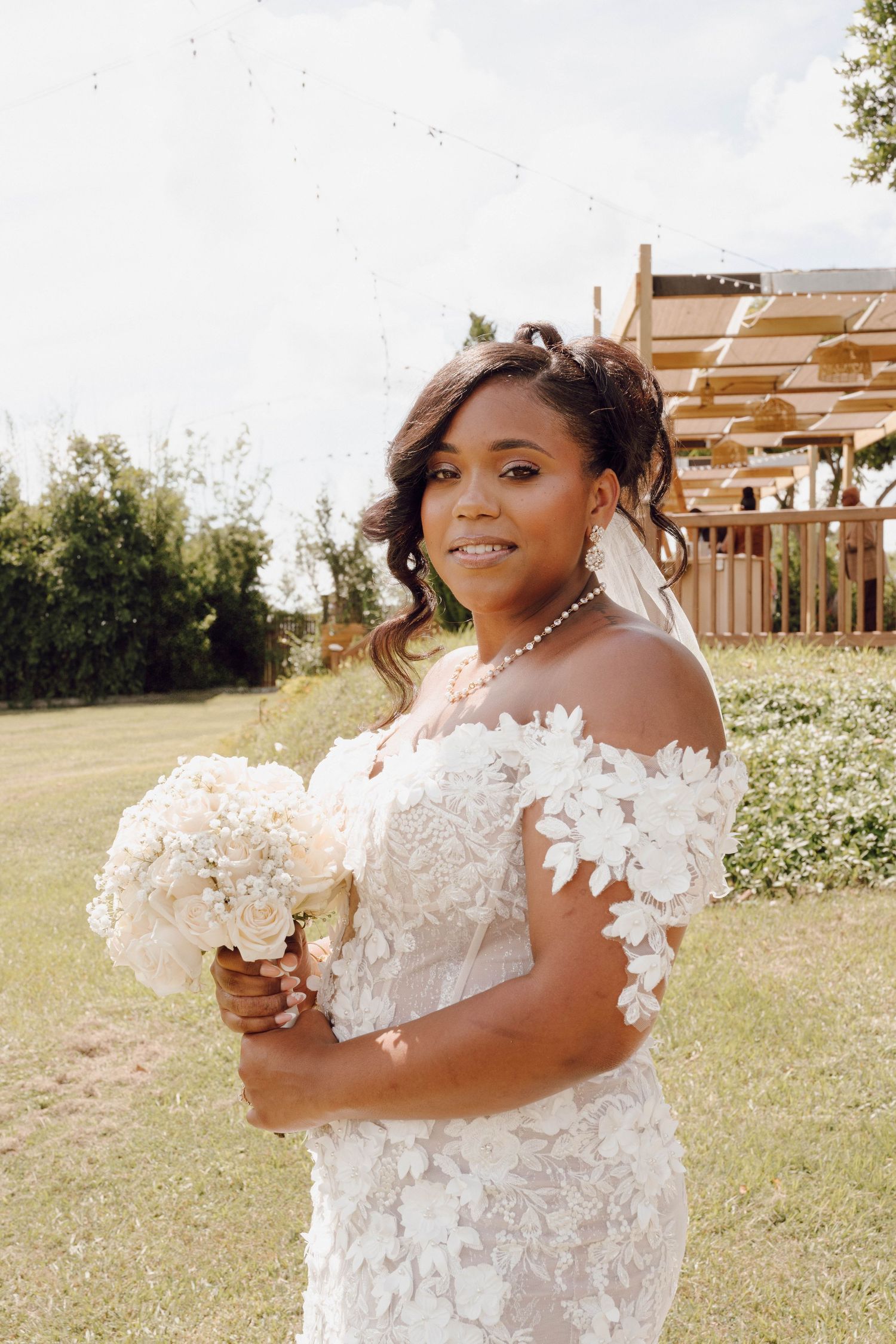 A bride in an elegant off-shoulder white lace wedding gown holds a bouquet while posing in a sunny outdoor garden setting.
