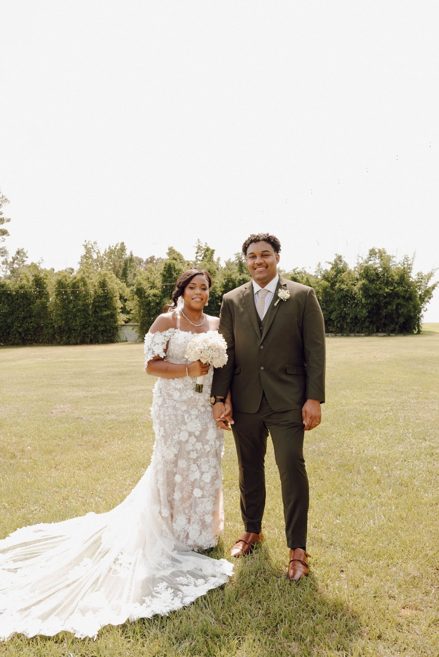 A newlywed couple poses outdoors on their wedding day with a beautiful lace dress and brown suit.