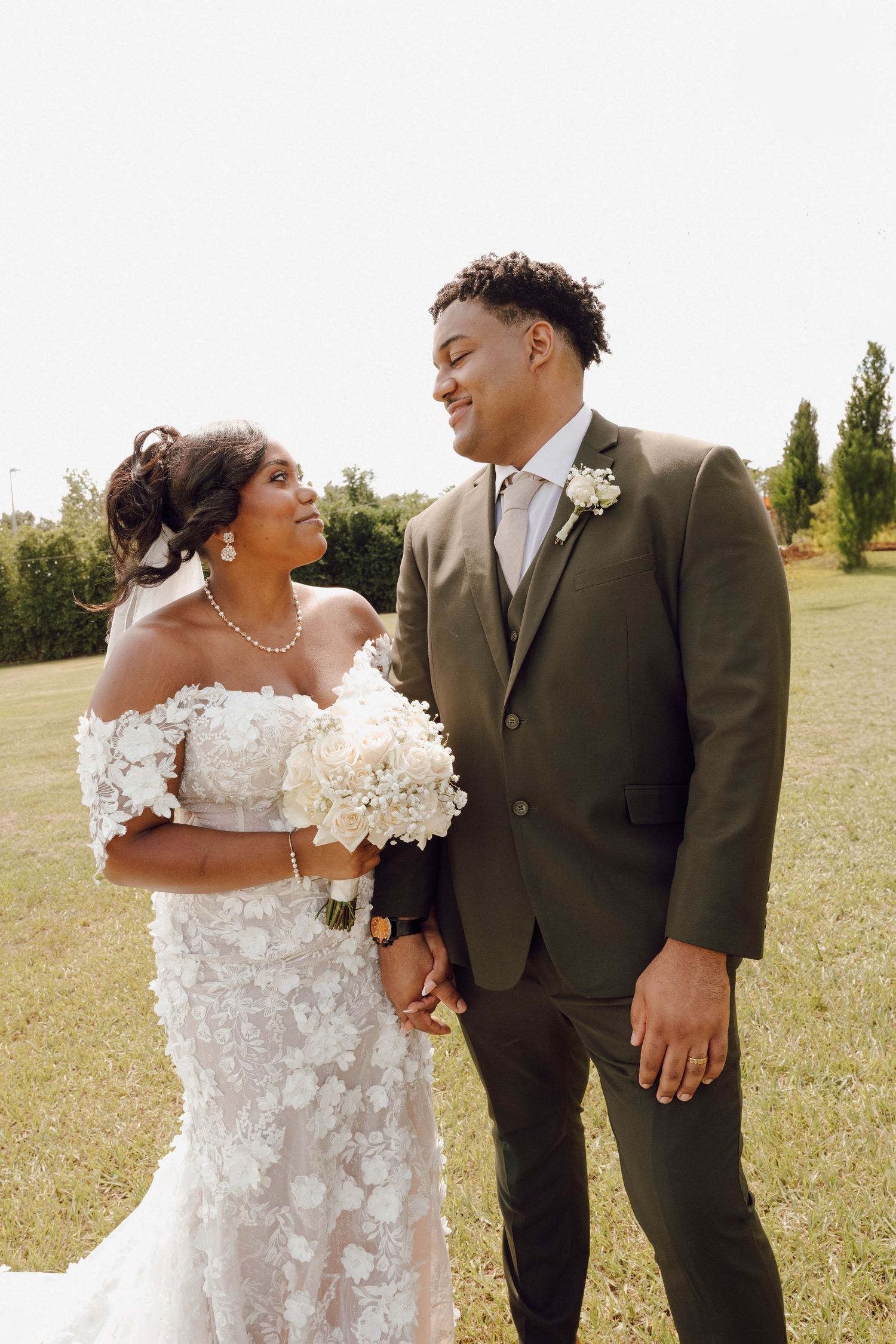 A joyful wedding couple shares a loving moment outdoors, with the bride in a floral lace gown holding white flowers.