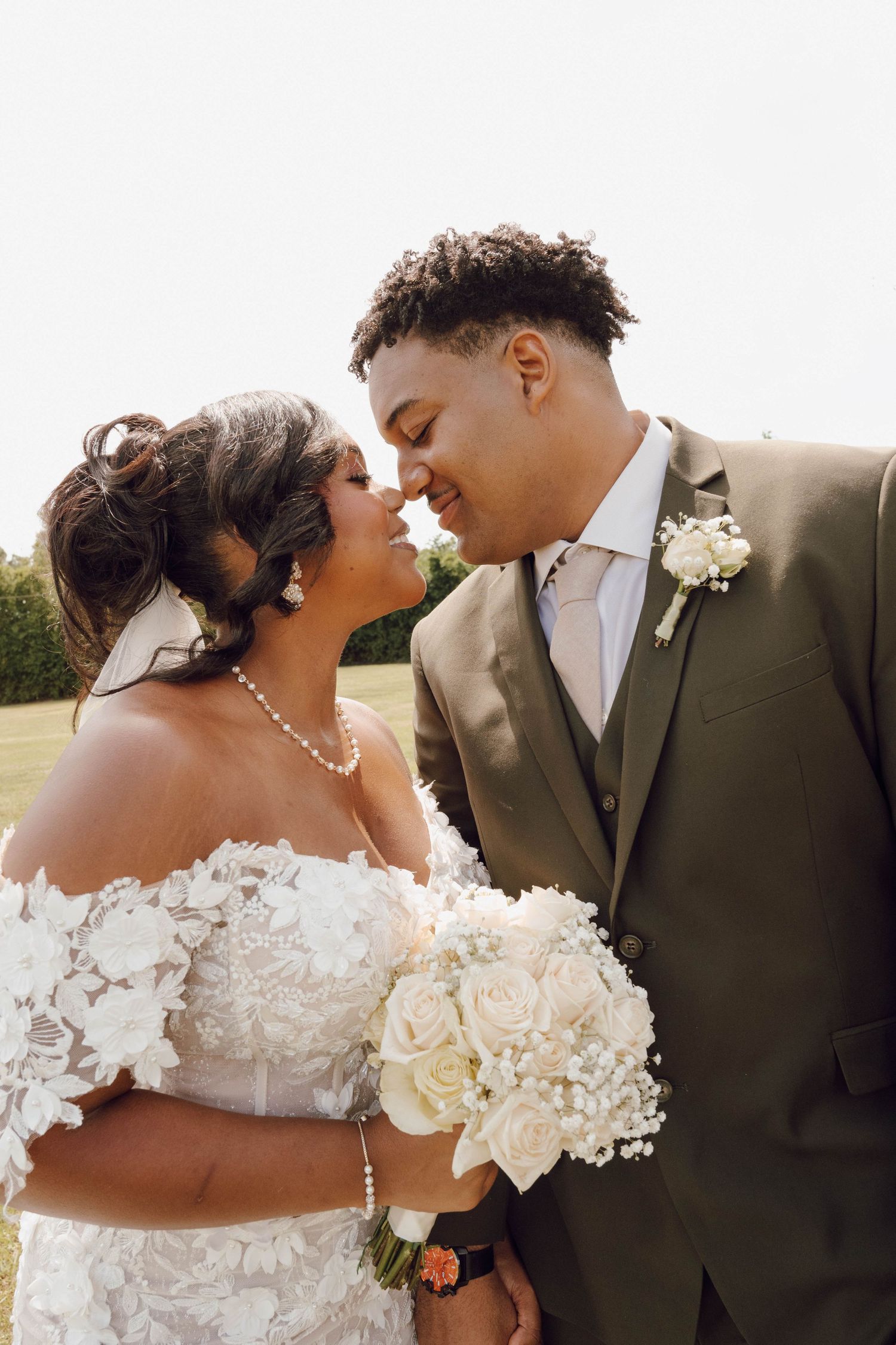 A romantic wedding portrait captures a tender moment between newlyweds against a sunny outdoor backdrop.
