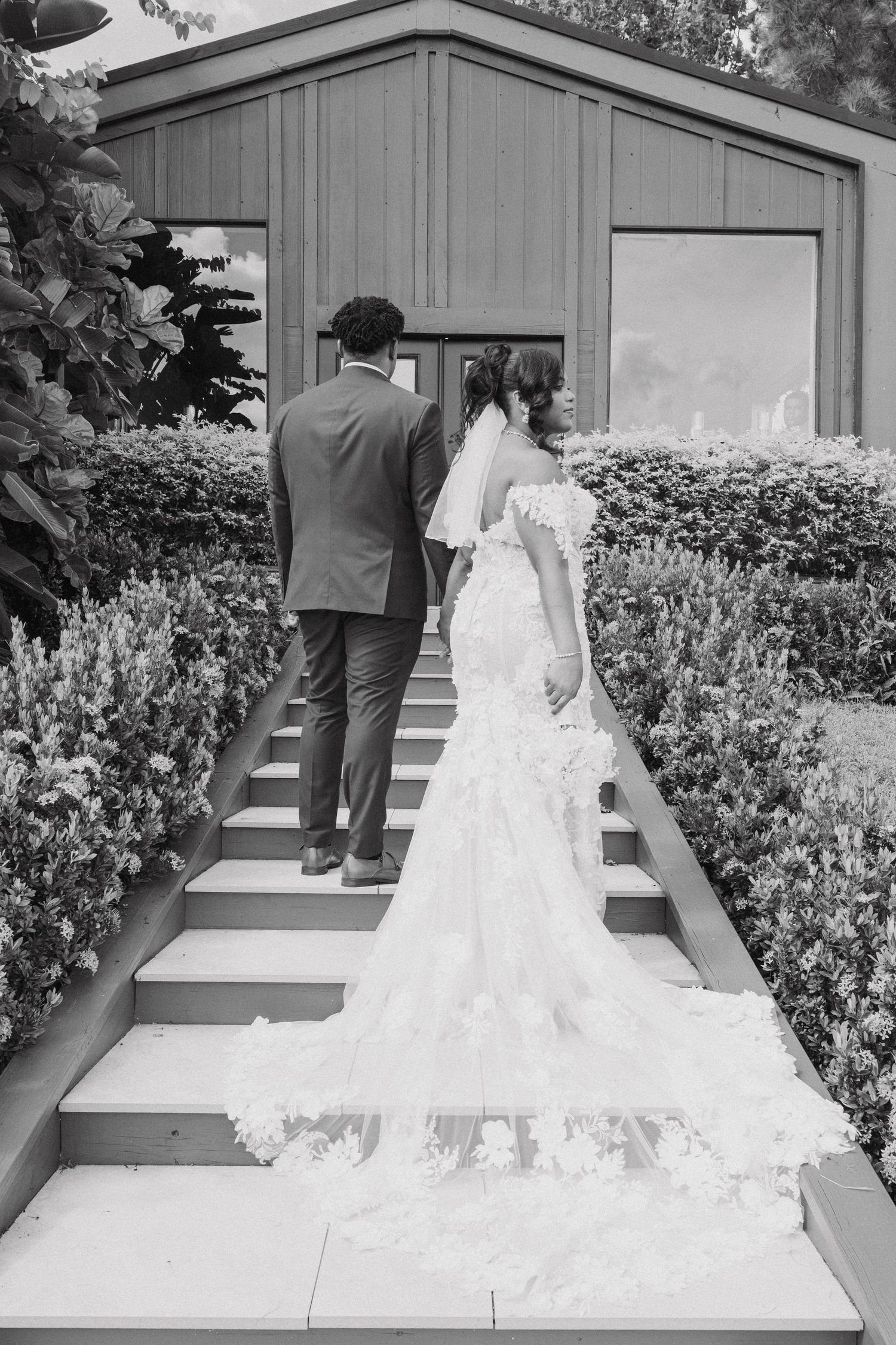 A black and white photo shows a bride's flowing train as the couple walks up garden steps.