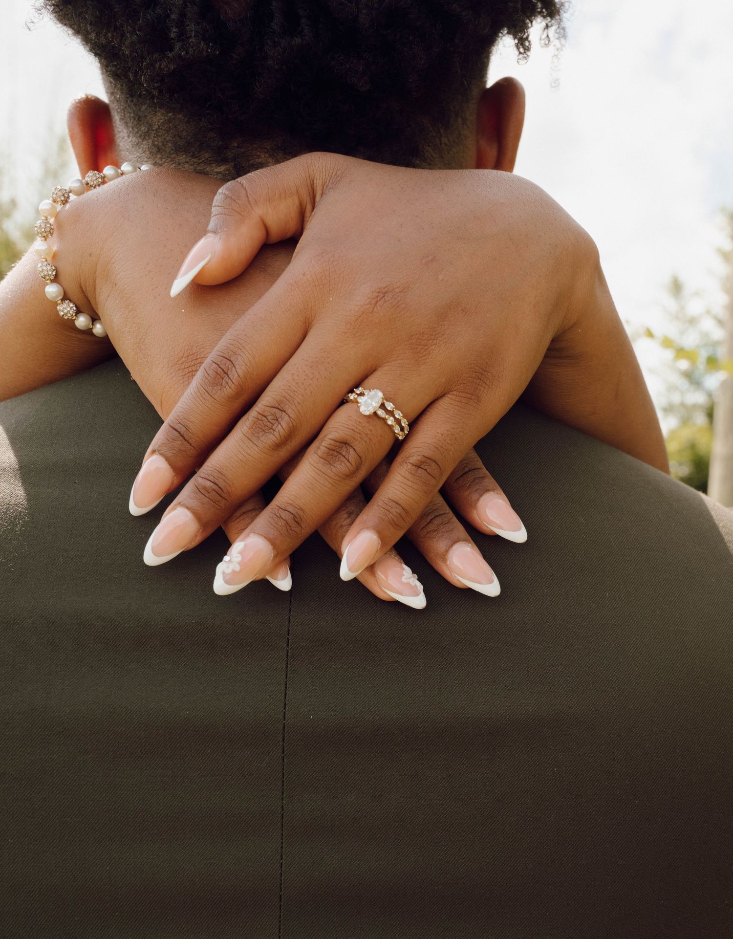 Close-up of hands with French manicured nails and engagement ring embracing in a romantic hug against olive green fabric.