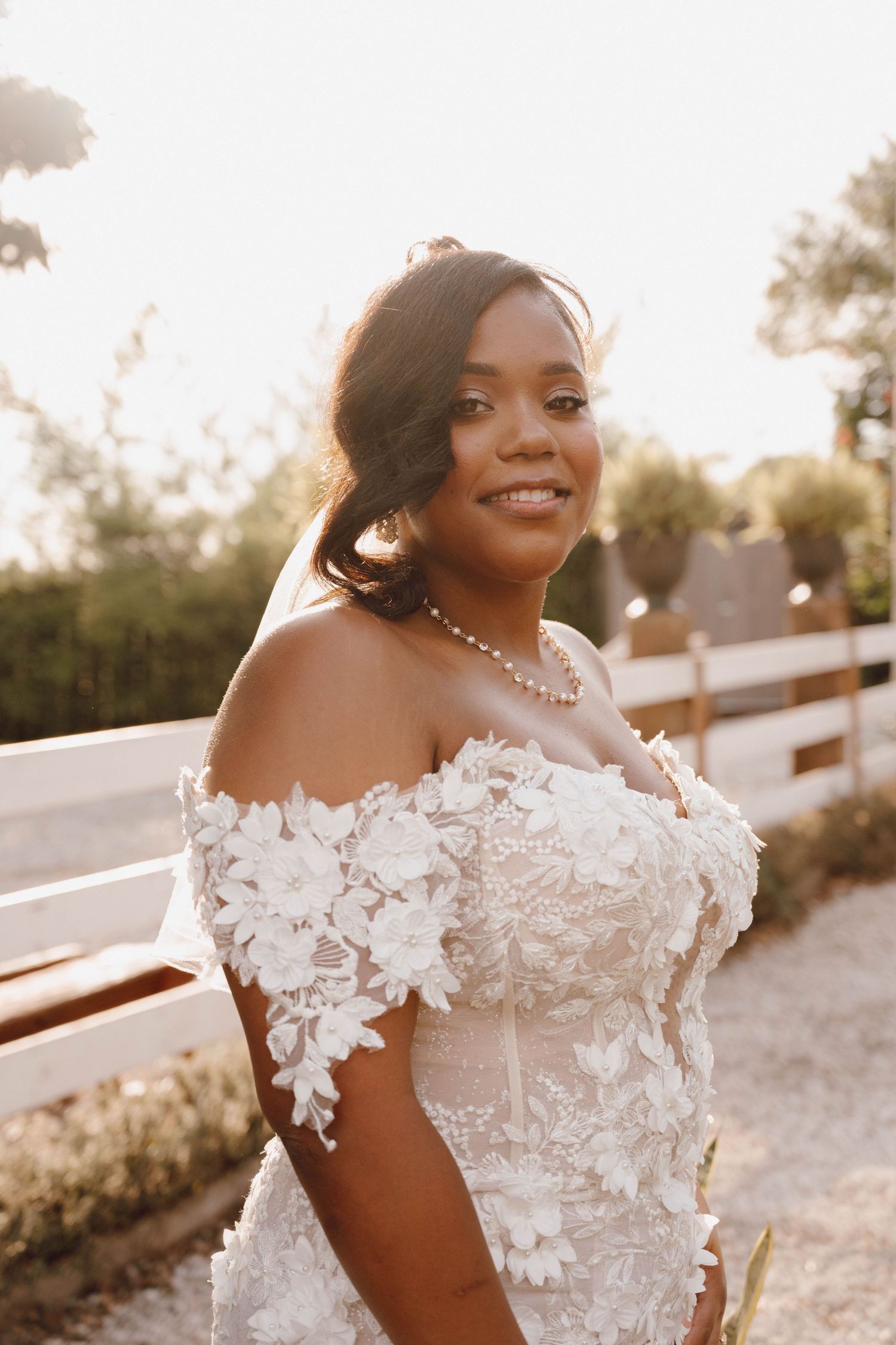Beautiful bride in an off-shoulder lace wedding gown with floral appliques poses during golden hour.