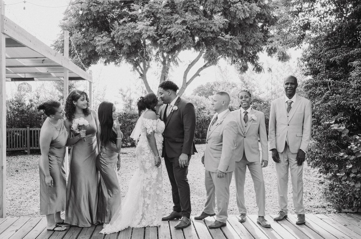 Wedding party gathered on a wooden deck under trees for formal group photos.