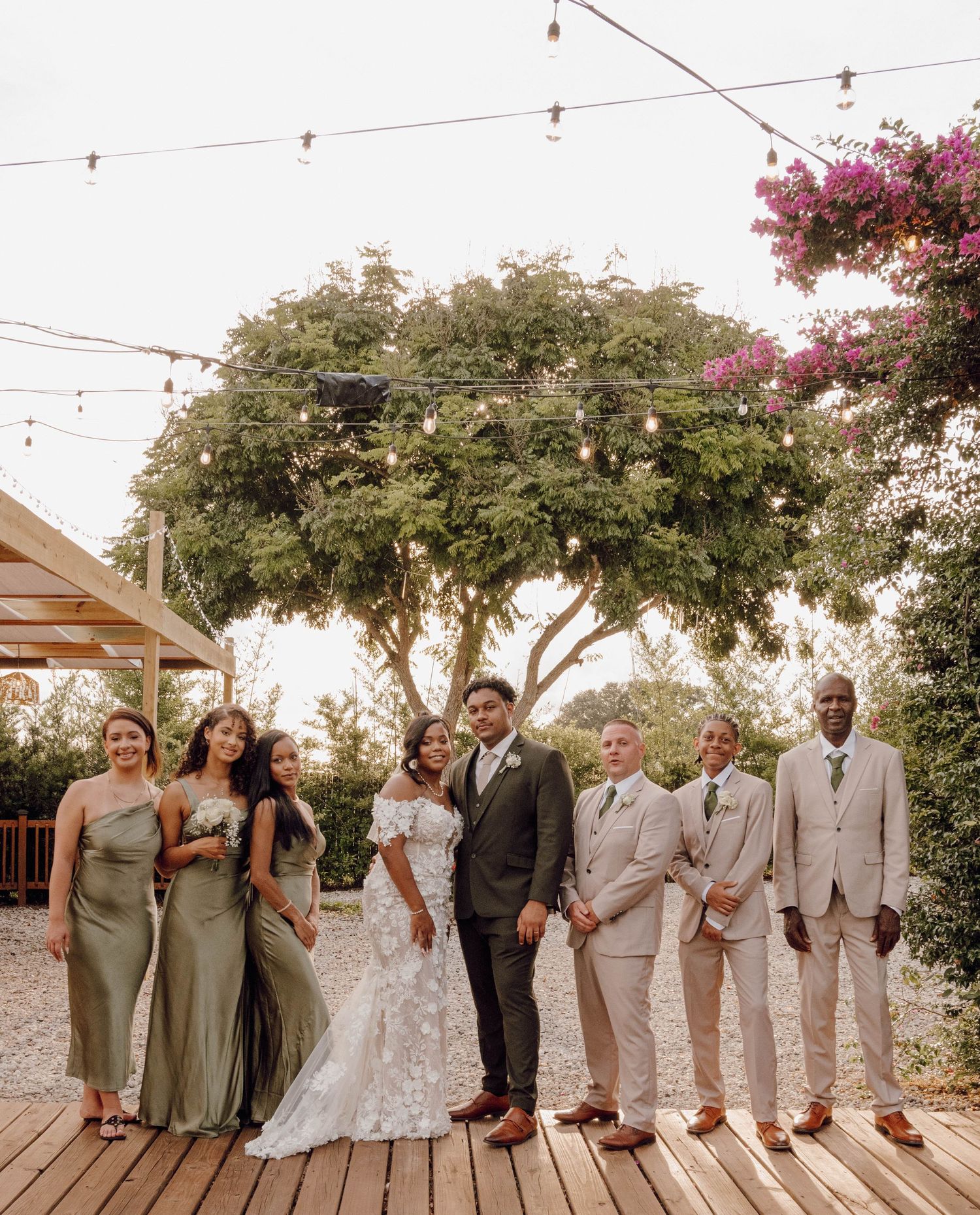 Wedding party poses together on a wooden deck with string lights and flowering trees in the background.