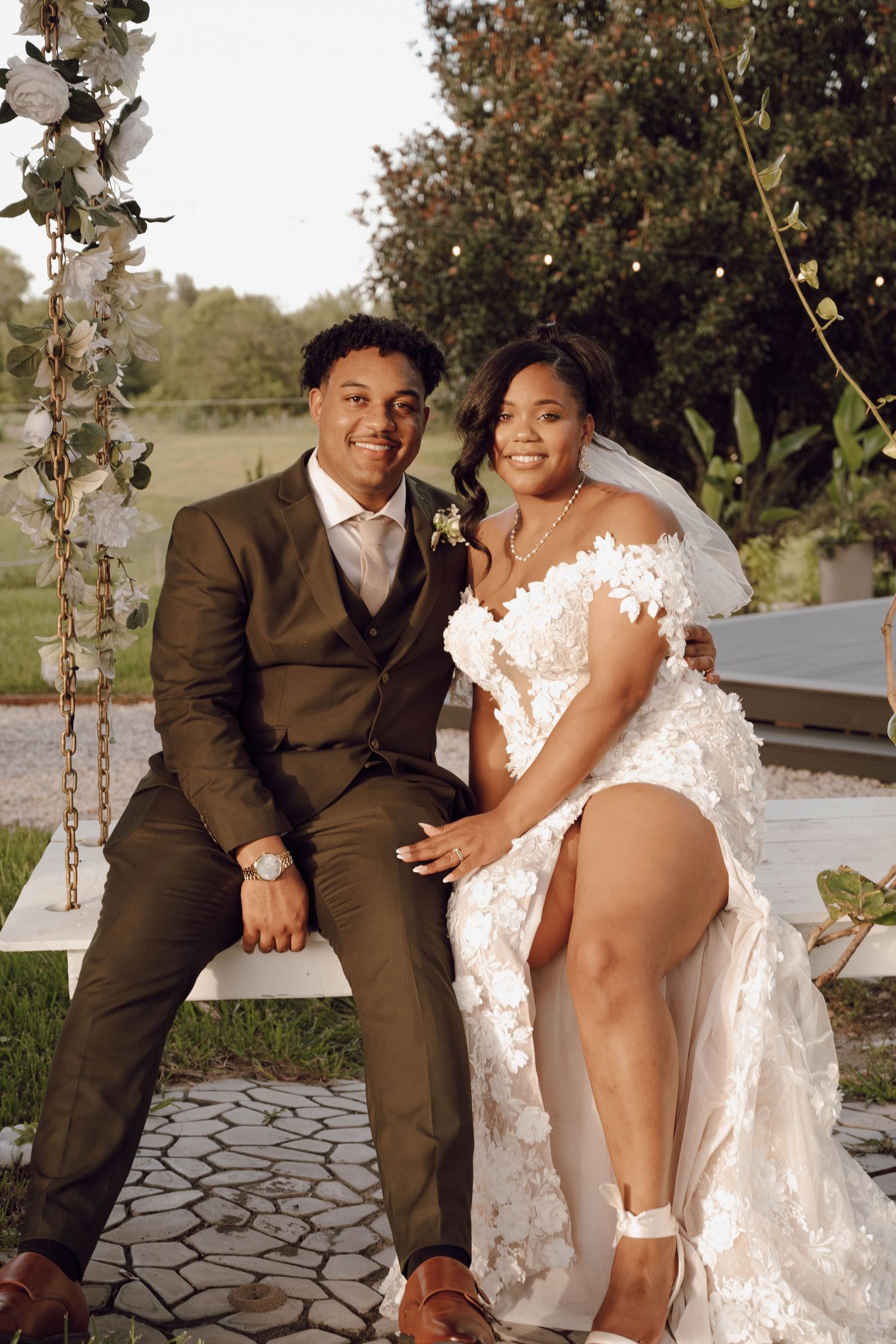 A romantic wedding photo of a couple sitting on a decorated swing in formal attire.