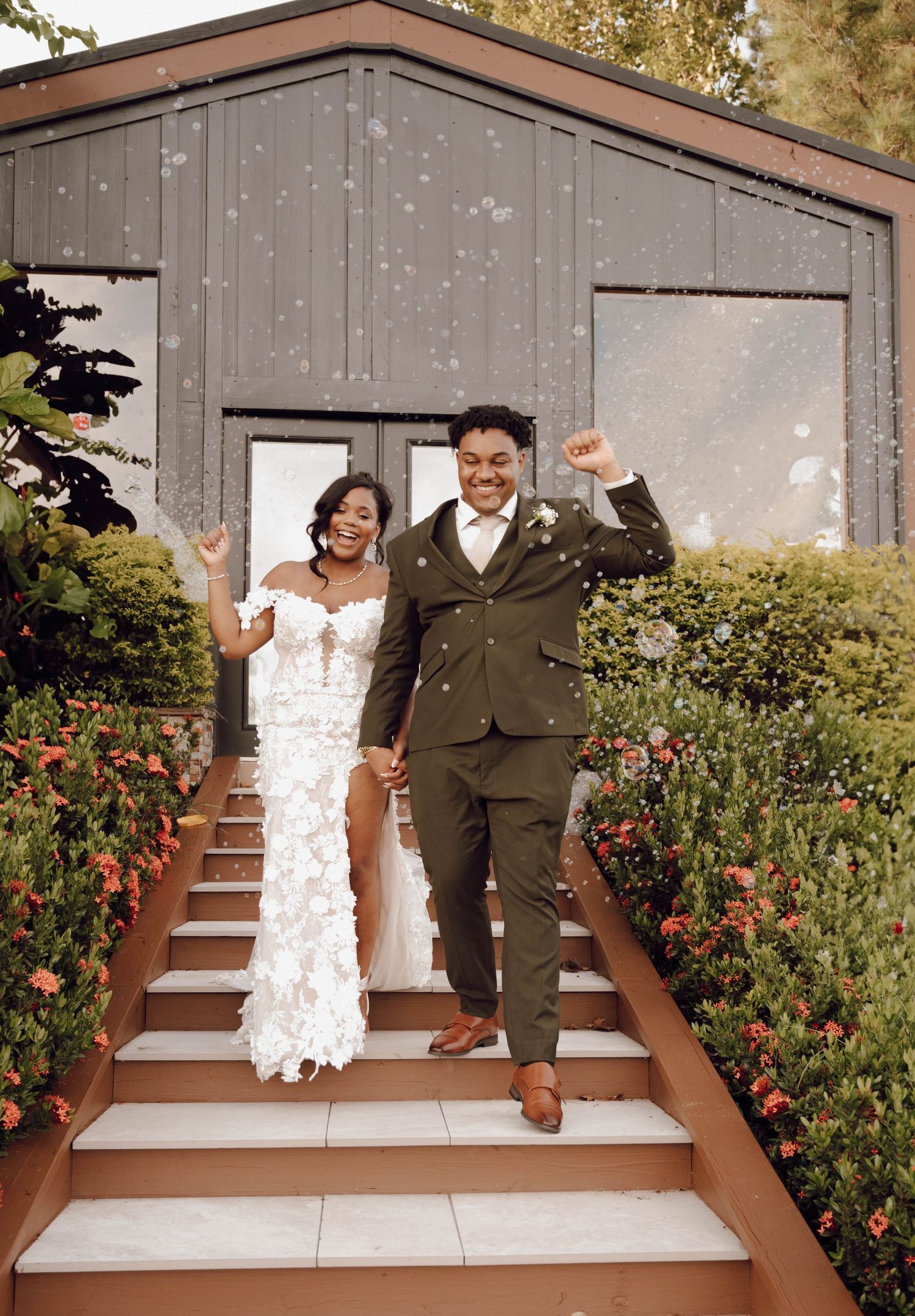 Newlyweds share playful jumping poses on outdoor steps surrounded by red flowers.